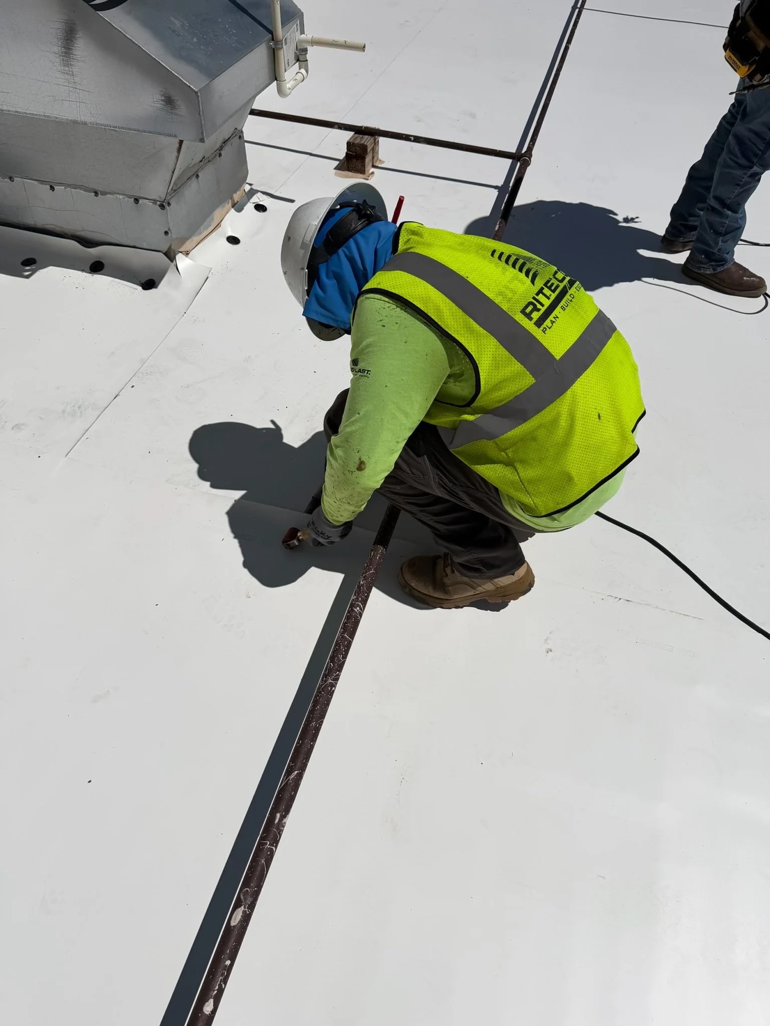 Worker wearing yellow safety vest and helmet installing insulation or ductwork on a white rooftop under bright sunlight.
