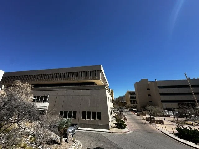View of modern office buildings under clear blue sky with minimal clouds, featuring trees and landscaping in the foreground.