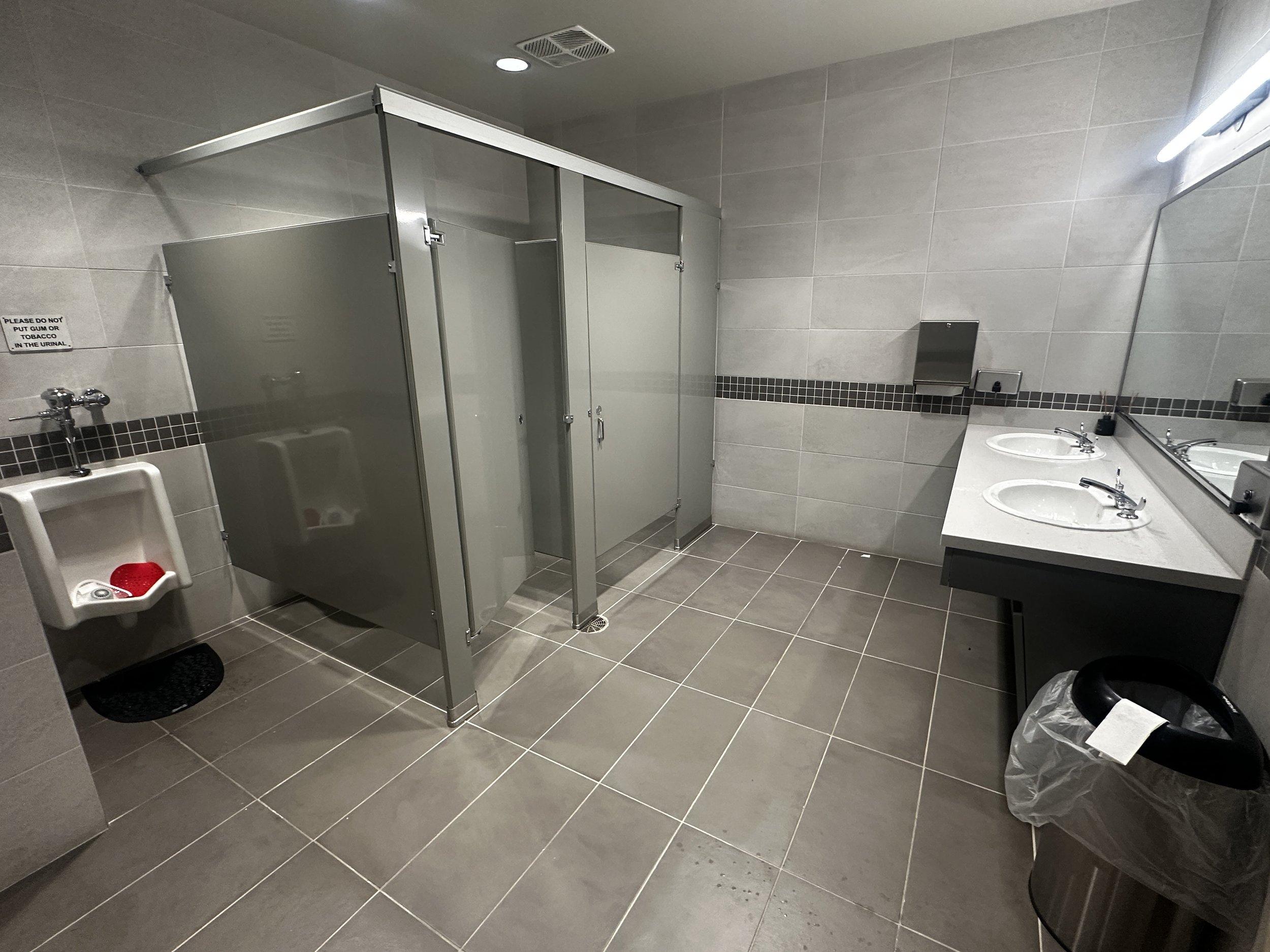 Public men's bathroom with gray tiled walls and floor, a row of sinks with stainless steel faucets, a mirror above the sinks, a soap dispenser, a paper towel dispenser, and a stall with a gray metal door.