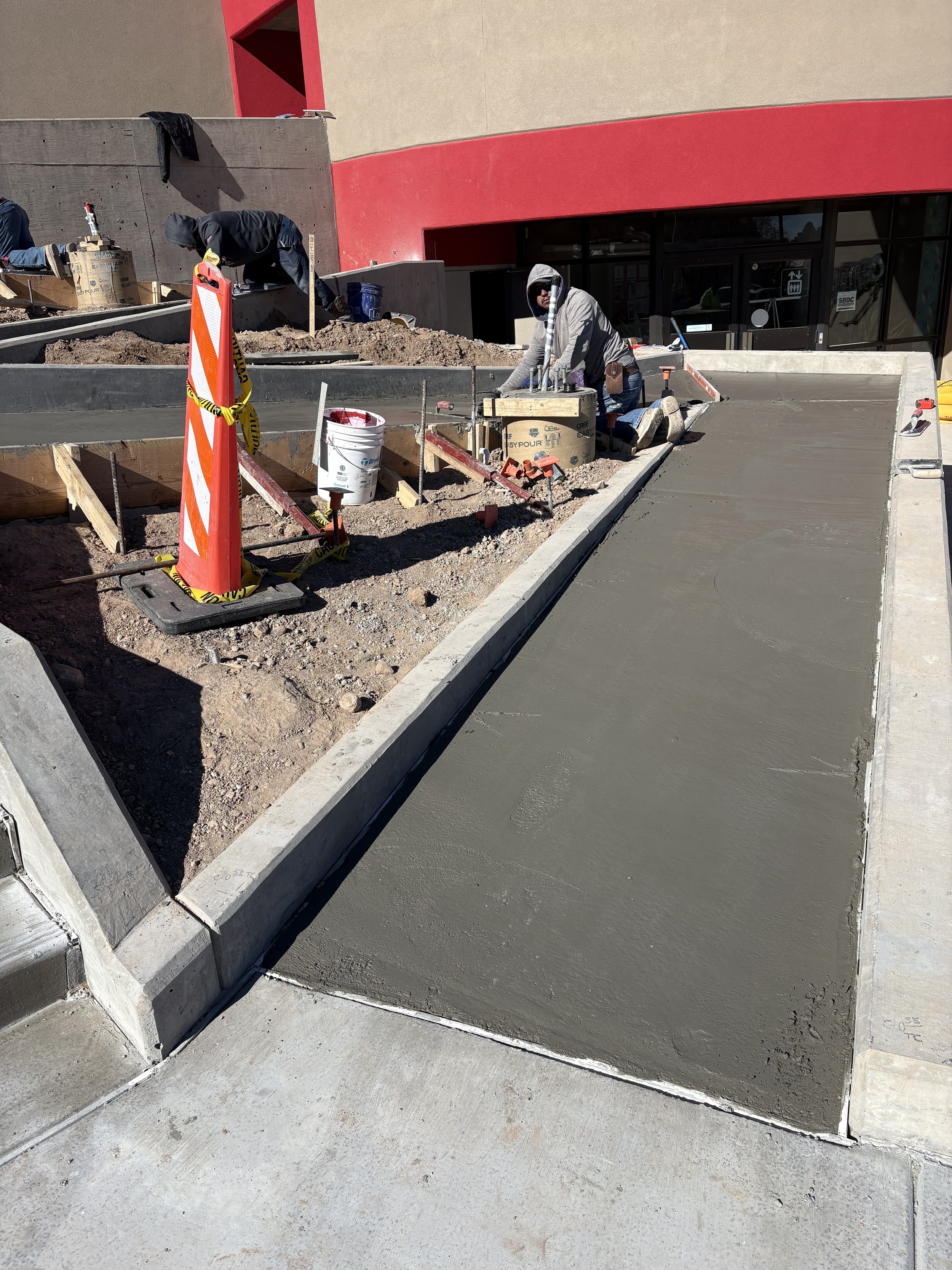 Construction workers laying fresh concrete on a sidewalk outside a building with a red and beige exterior.