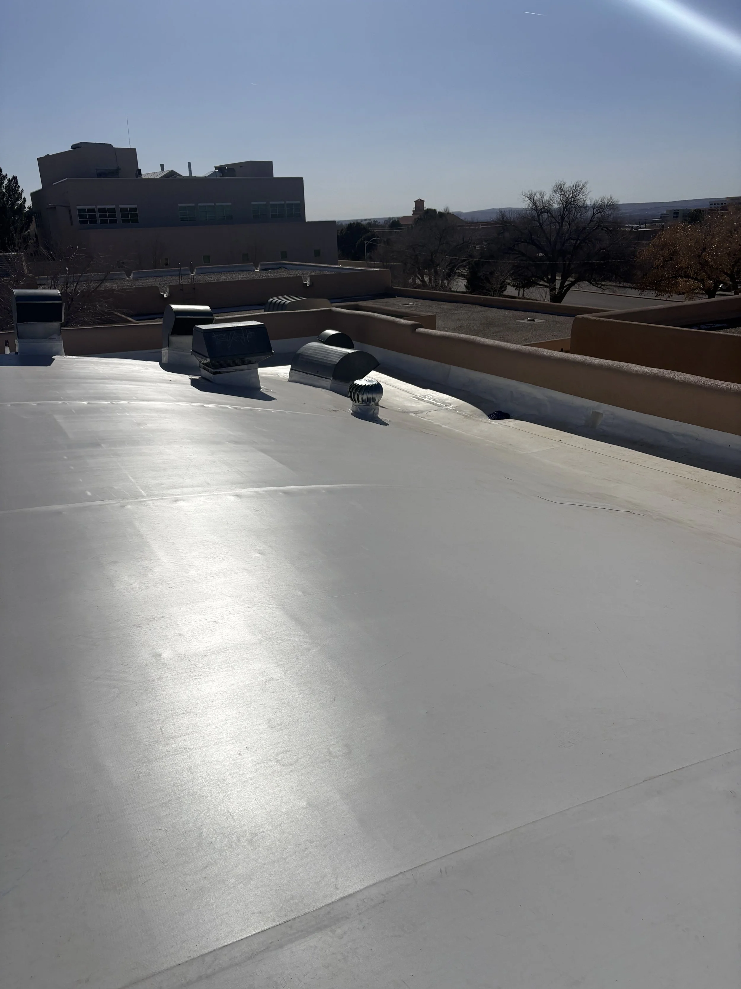 View of a flat rooftop with multiple ventilation ducts and a beige parapet wall, with leafless trees and a large building in the background under a clear sky.