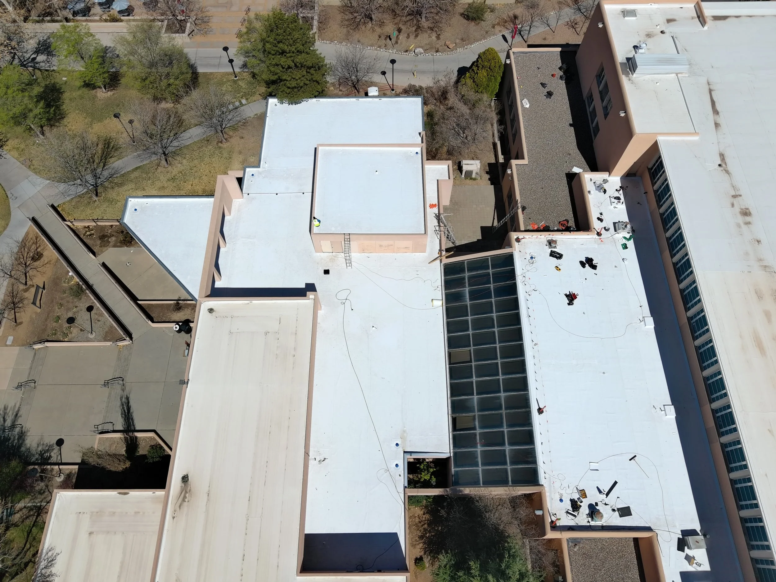 Aerial view of a multi-story building with flat white roofs, some with equipment, outdoor courtyard, trees, and surrounding sidewalks.