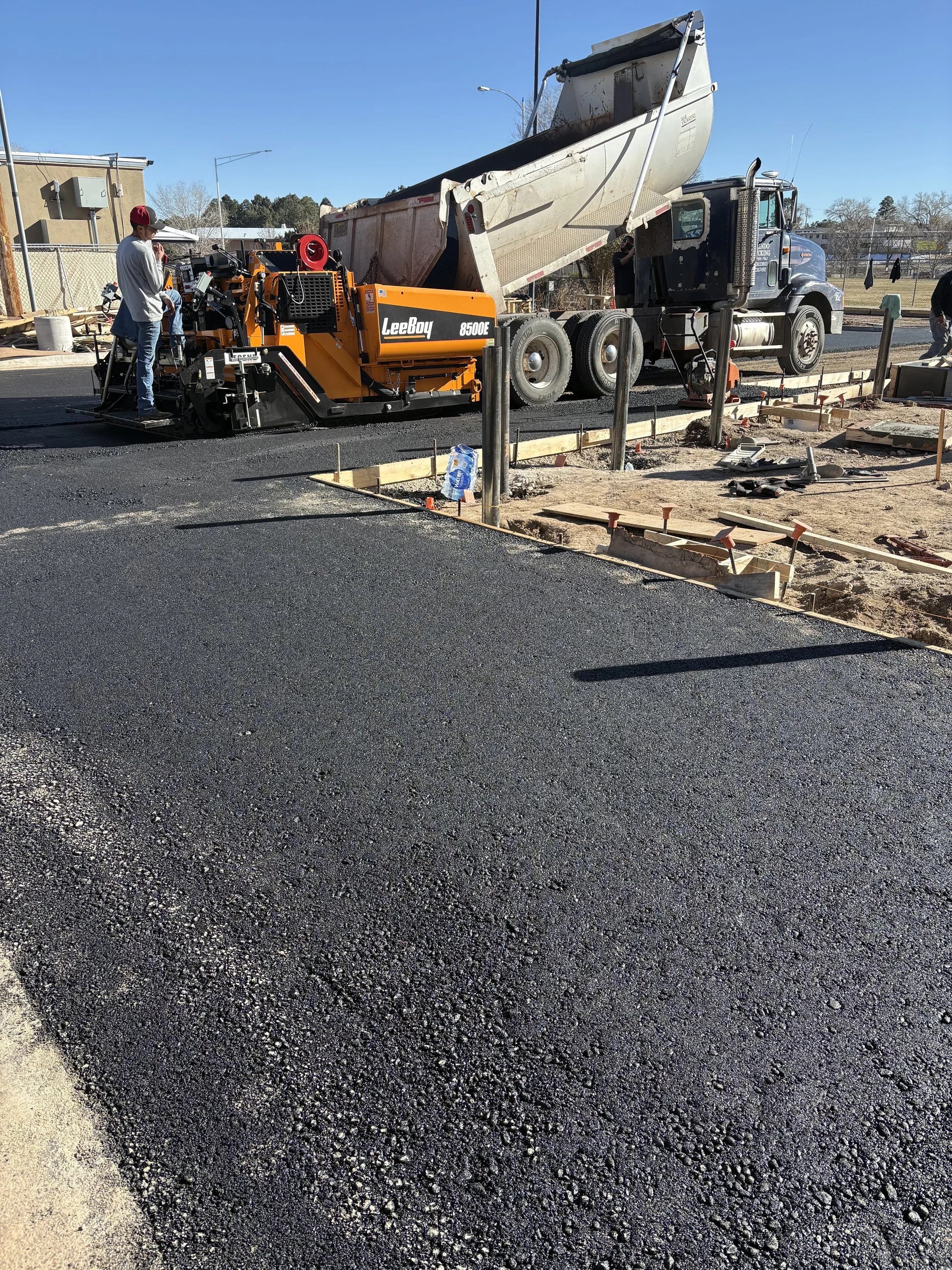 Construction workers are paving asphalt on a road. A large dump truck is pouring asphalt, and a paving machine is spreading it evenly. Construction tools and materials are scattered on the ground.