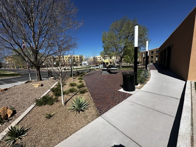 Empty outdoor plaza with trees, benches, trash cans, a brick pathway, and surrounding buildings under a clear blue sky.