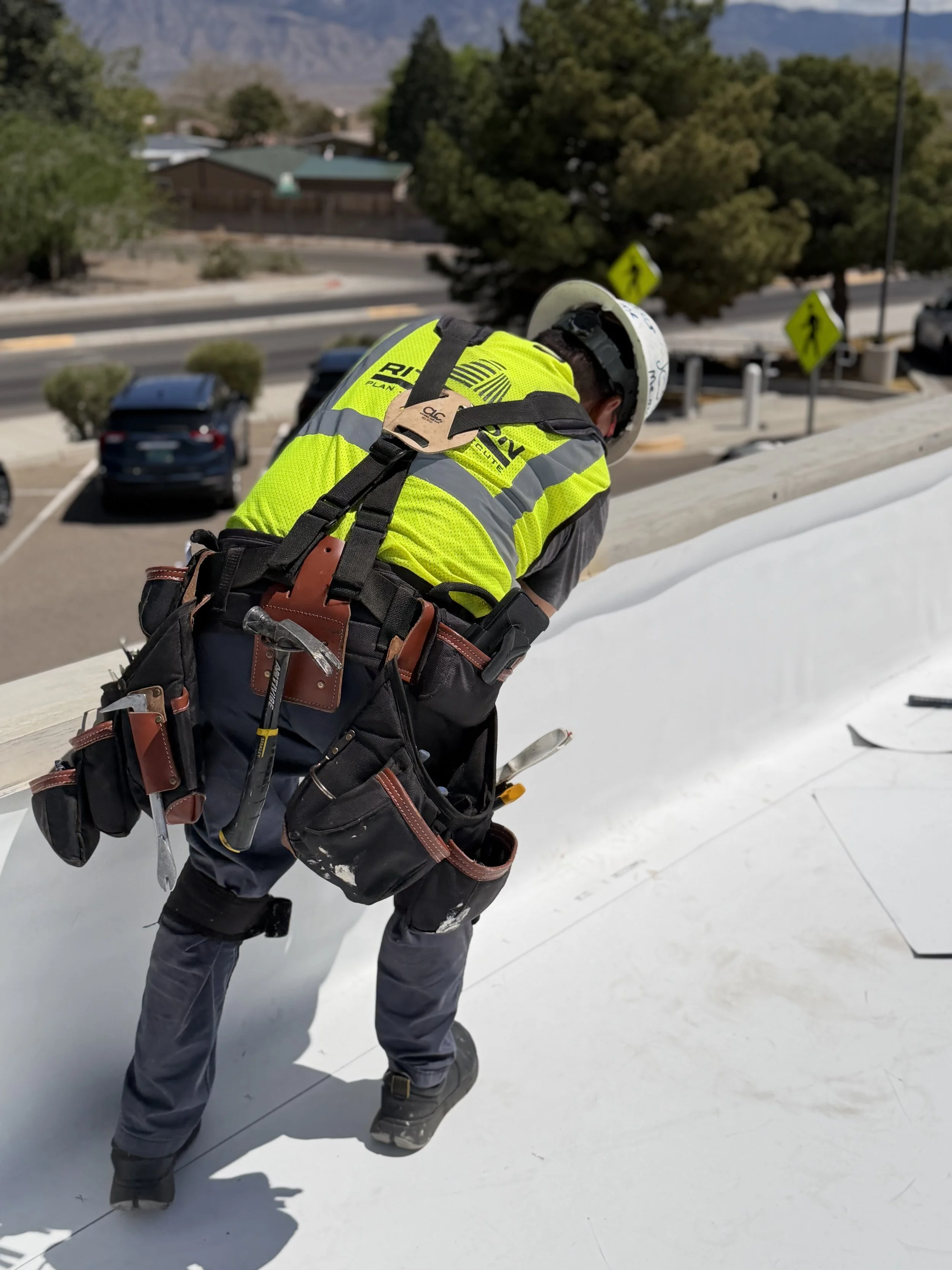 A construction worker wearing a yellow safety vest, a white helmet, and tools around his waist, working on a white roof in a residential area with trees, parked cars, and a street in the background.