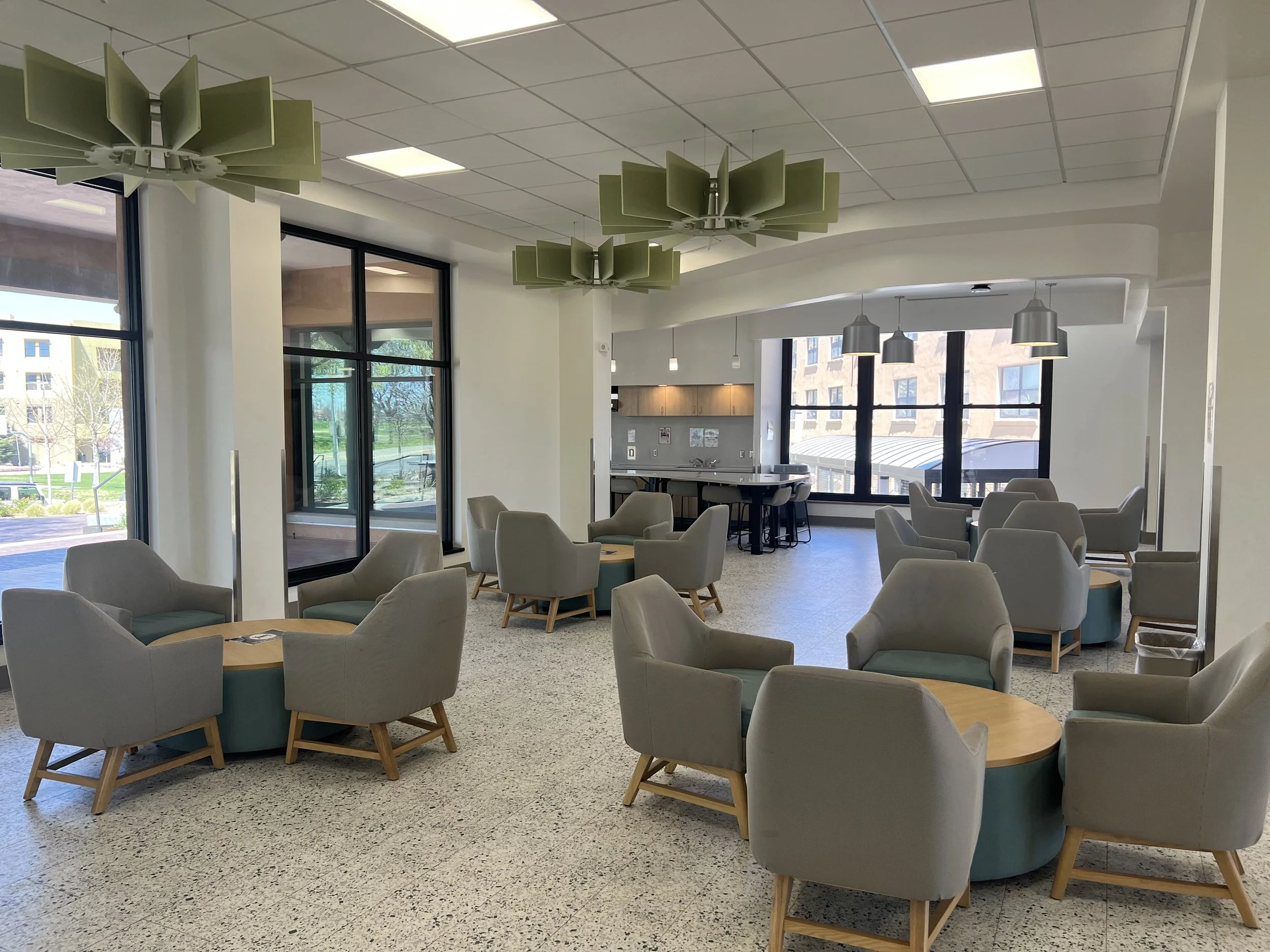 Interior of a modern lounge or cafeteria with large windows, multiple seating arrangements of armchairs and small tables, hanging green and gray ceiling lights, and a partial view of a kitchen area.