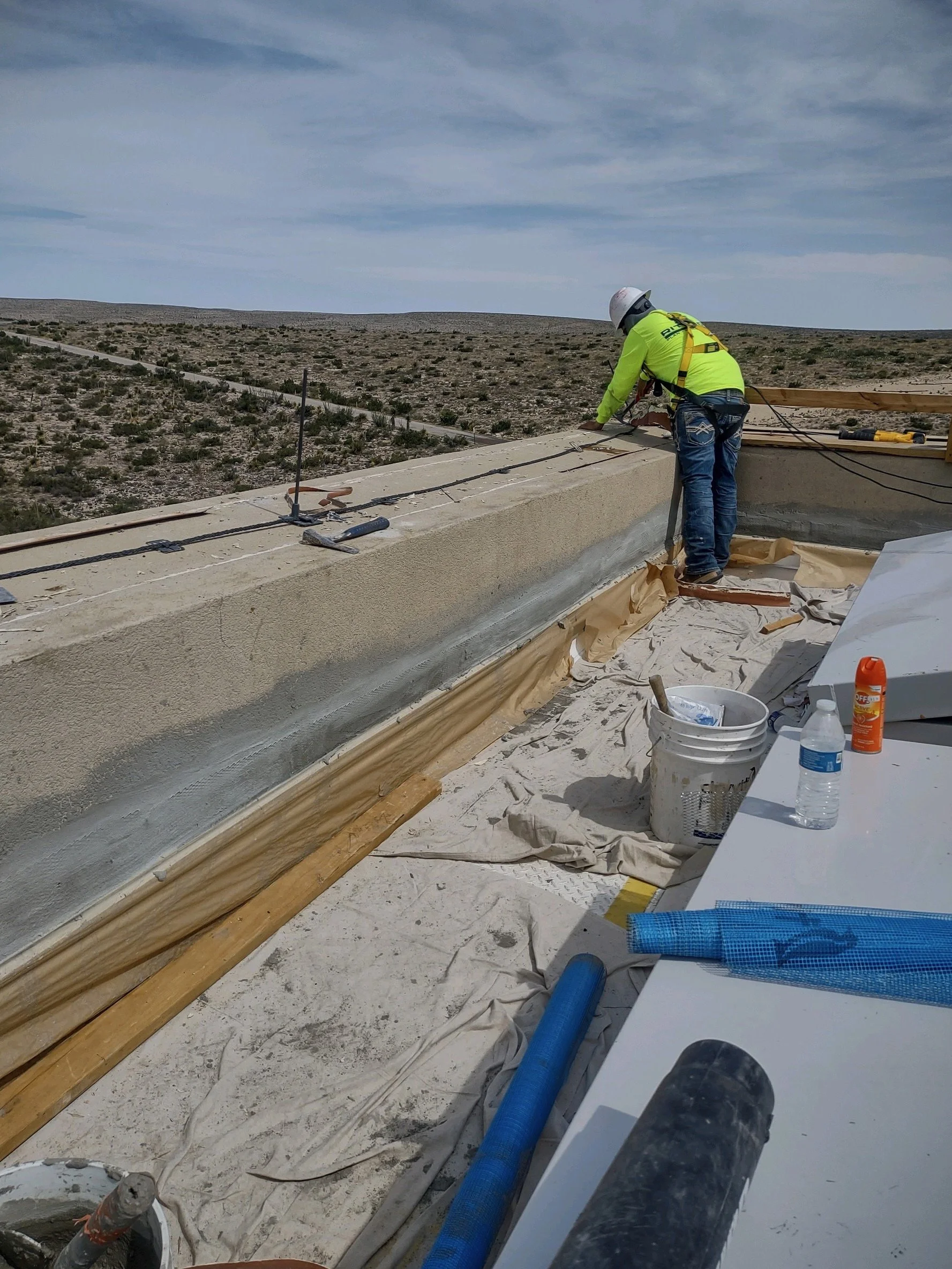 A construction worker in a yellow safety shirt and jeans is working on the edge of a building's rooftop. The worker is wearing a white helmet and a safety harness while securing cables or wires on a parapet wall. The background shows a barren landsca