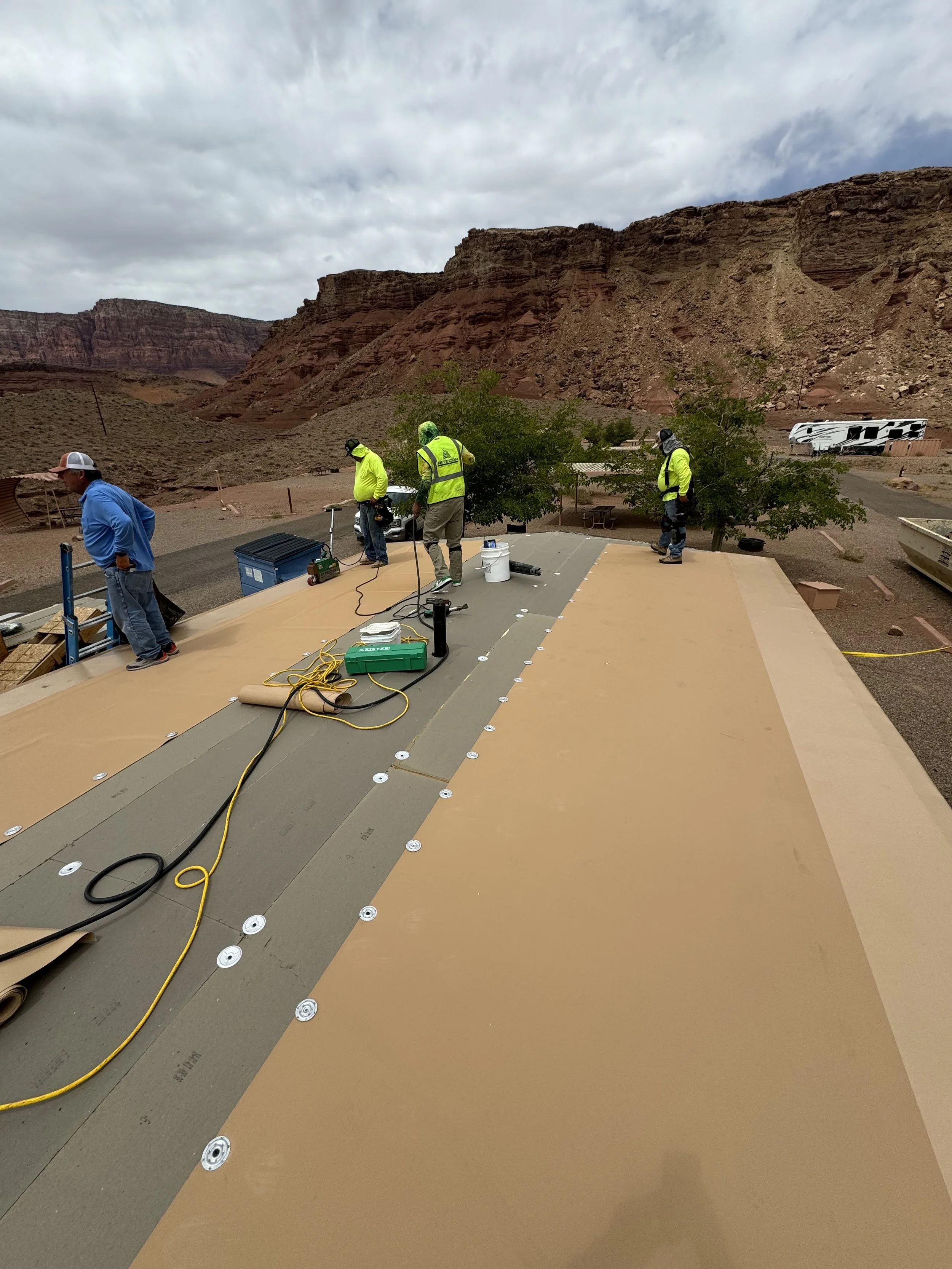 Construction workers installing insulation panels on a roof in a desert landscape with rocky cliffs in the background.