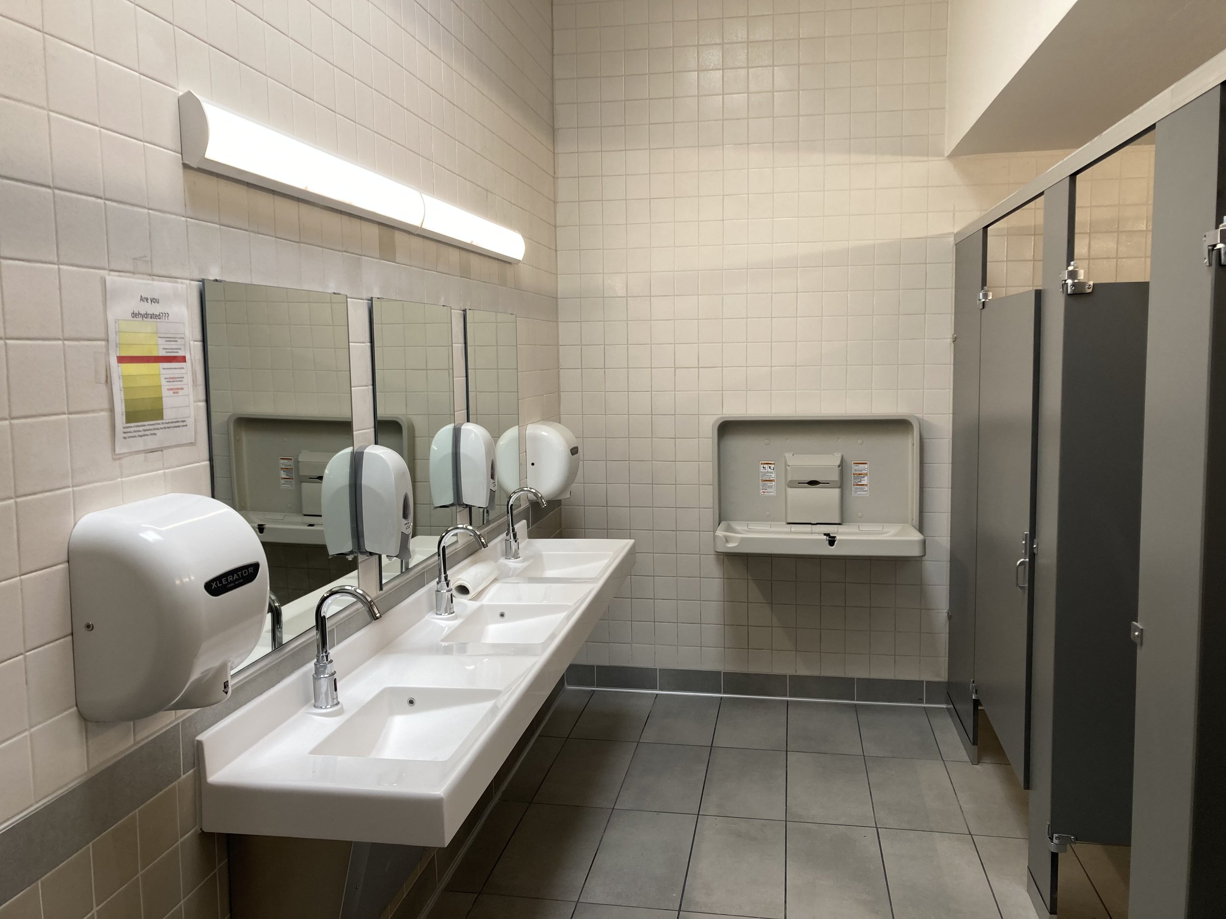 Public restroom with three sinks, mirrors, and hand dryers, beige tiled walls, gray floor, and partially visible toilet stall.