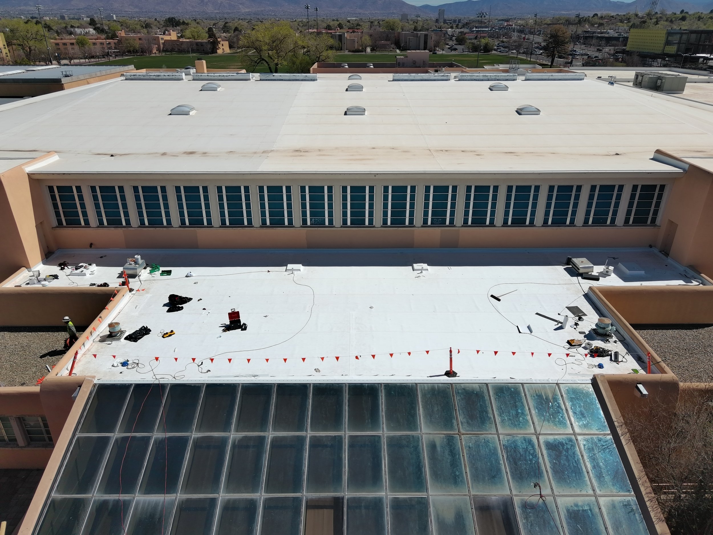 Aerial view of a building roof with vents and equipment under repair, with neighboring buildings and trees in the background.