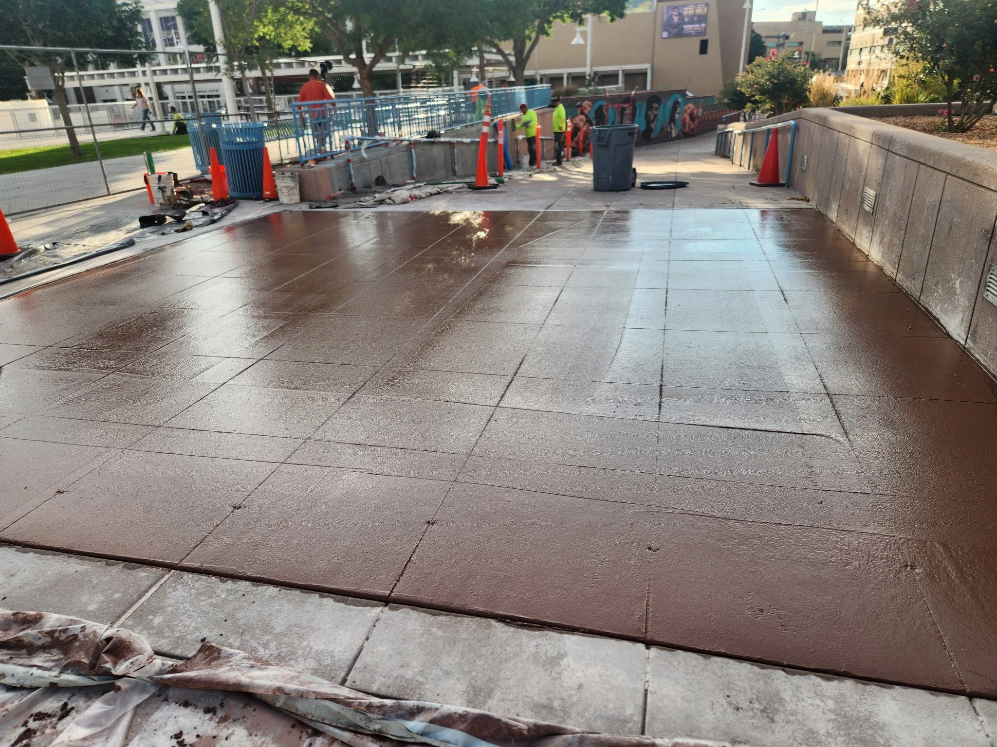 Construction workers are laying down new brown tiles on the sidewalk in an urban area, with construction cones and equipment nearby.