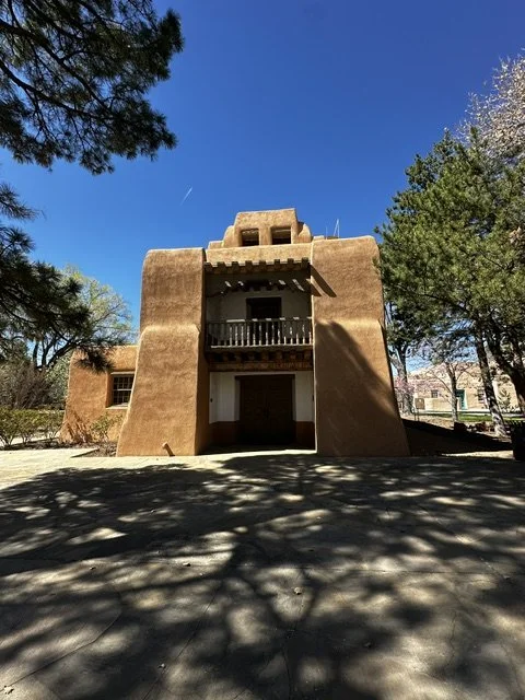 A historic adobe-style building with a balcony, surrounded by tall trees, under a clear blue sky.