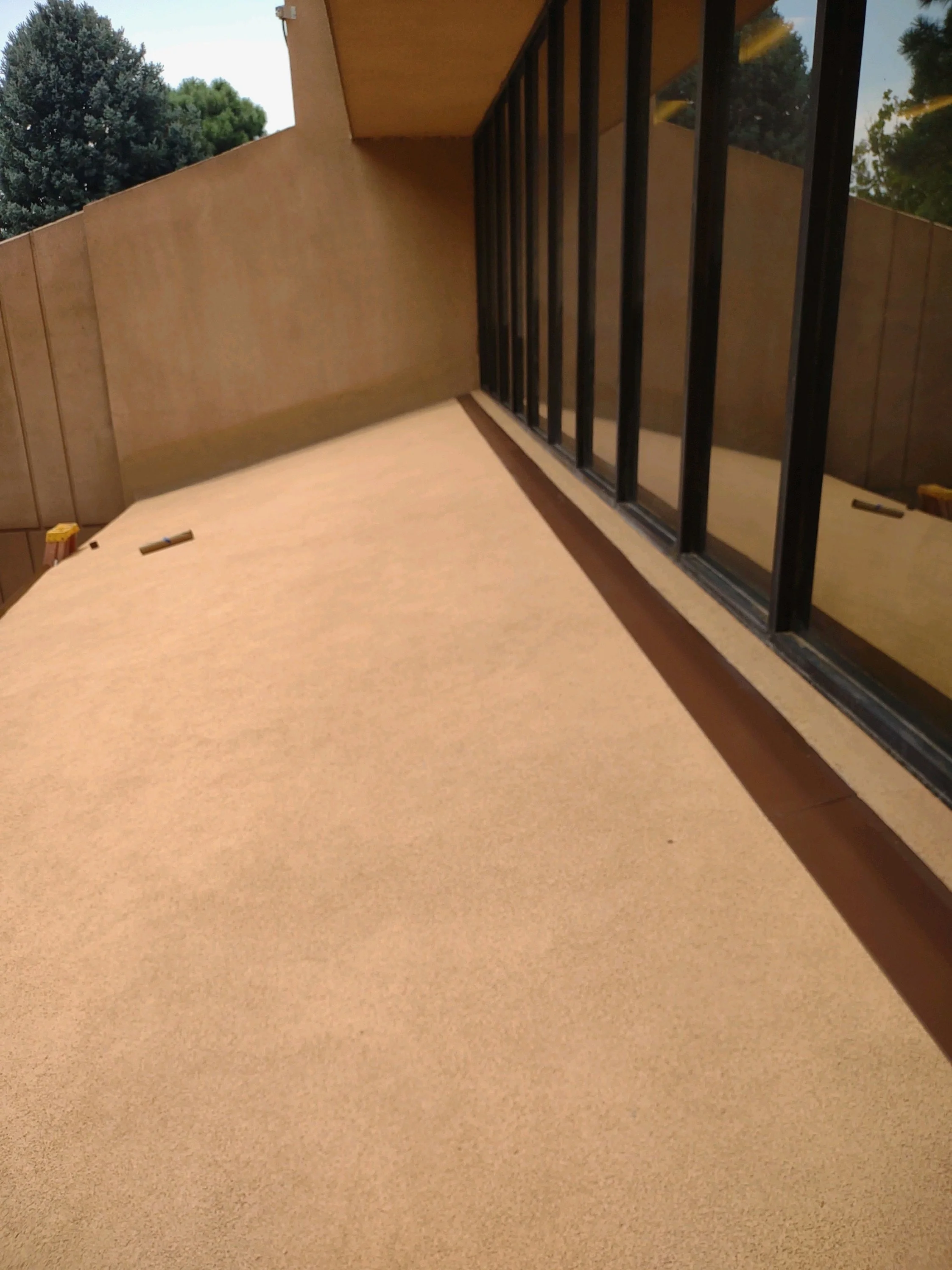 An outdoor balcony with beige and brown walls, a glass sliding door with black frame, and trees visible in the background.