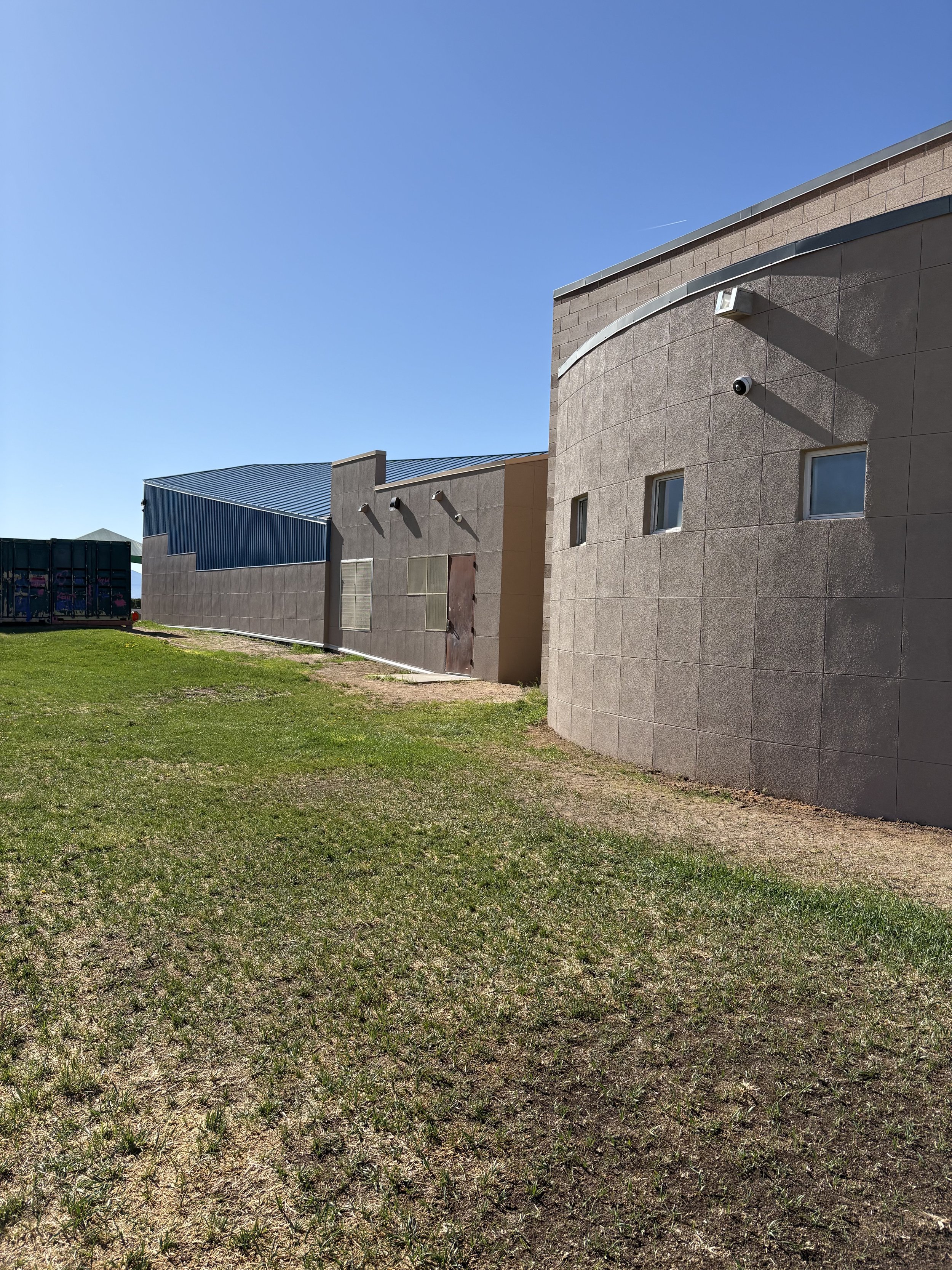 A building with a curved exterior wall, small windows, and external security cameras, under a clear blue sky with a patch of grass and dirt in the foreground.