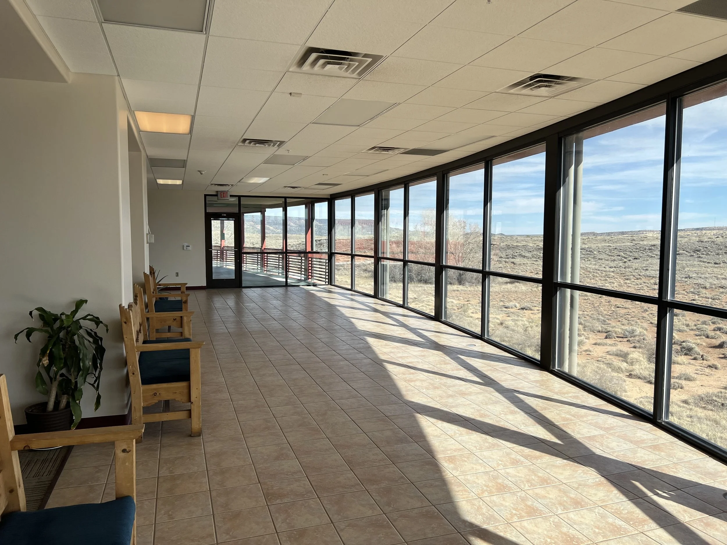 Interior of a room with large windows showing a desert landscape outside, wooden chairs lined along one side, and a potted plant near the wall.