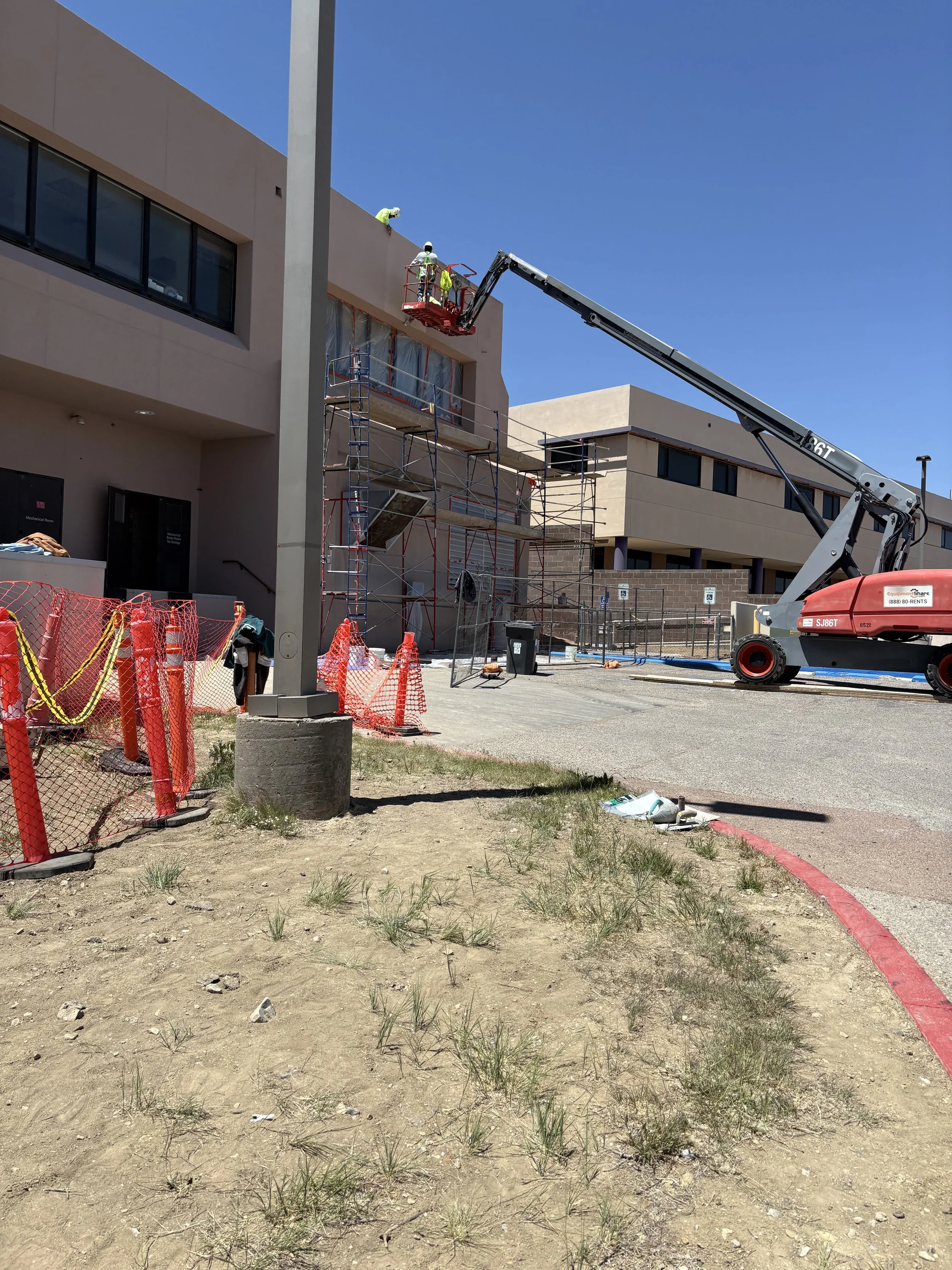 Construction workers using a cherry picker to work on the upper part of a modern building, with scaffolding and construction materials around, under a clear blue sky.