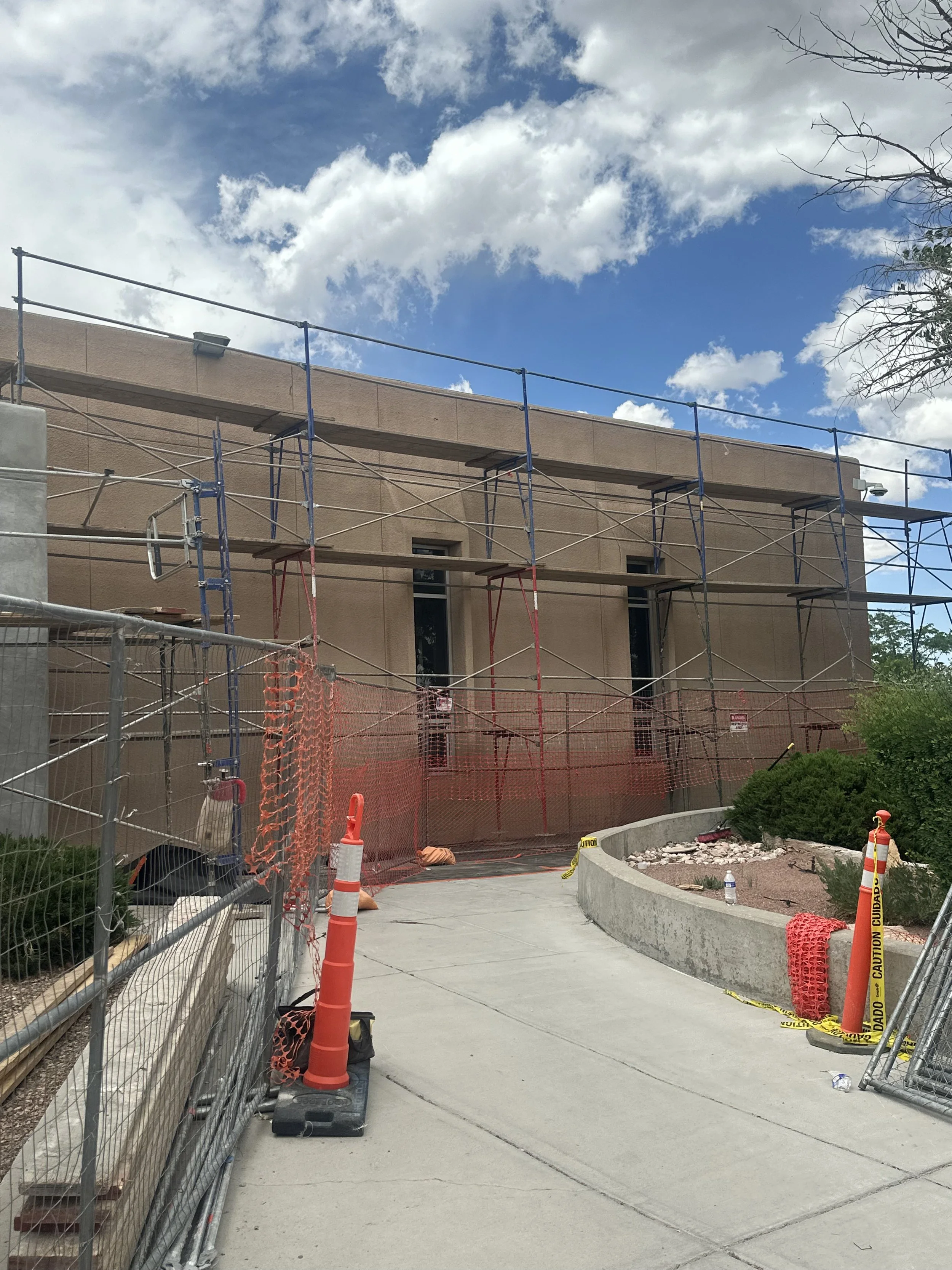 Construction site with scaffolding and orange safety barriers in front of a tan building with windows, under a partly cloudy sky.