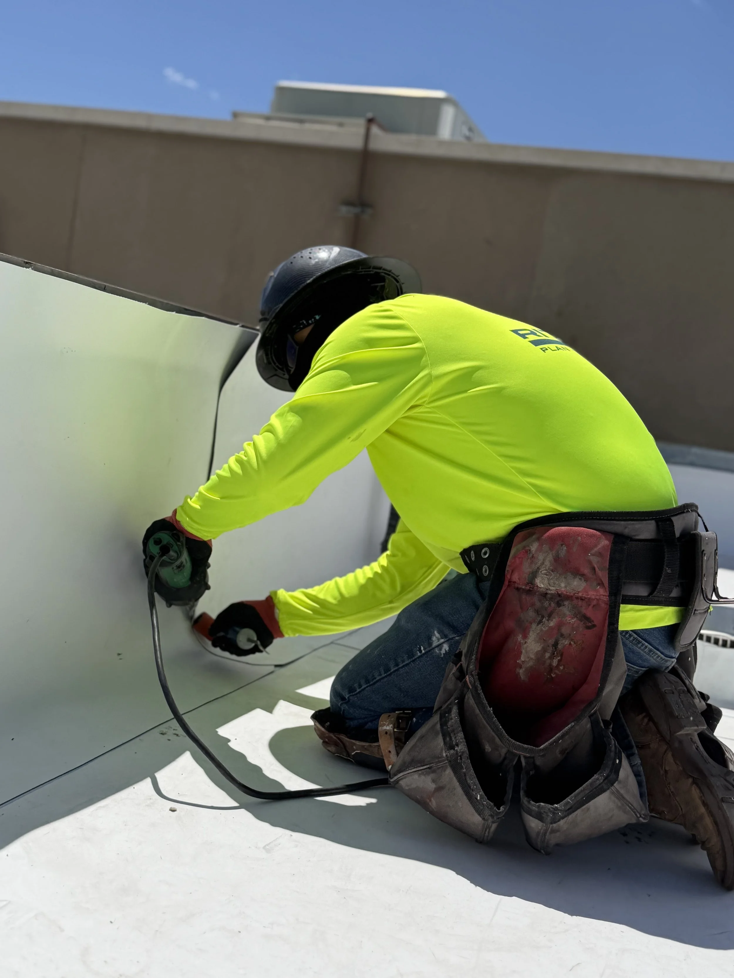 A worker in a bright yellow safety shirt kneels on a white rooftop, using a power tool on the surface. The worker is wearing a black helmet and a tool belt. The sky is clear and blue.