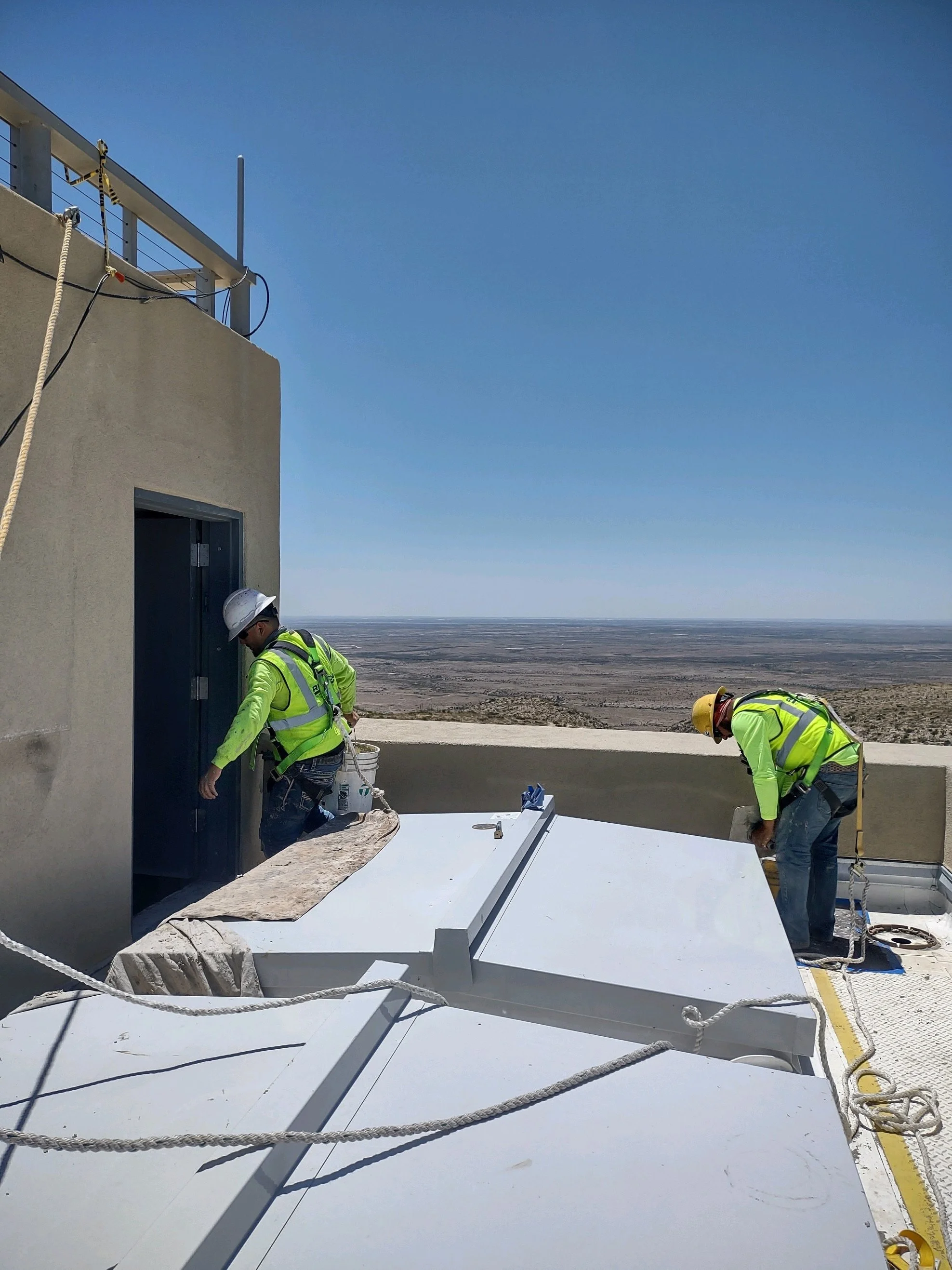 Two construction workers in safety gear working on a rooftop during daytime, with a clear sky and an expansive desert landscape in the background.