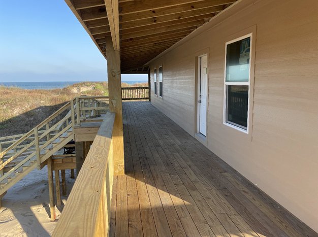 View of a wooden balcony attached to a house overlooking a beach with dunes and water in the distance.