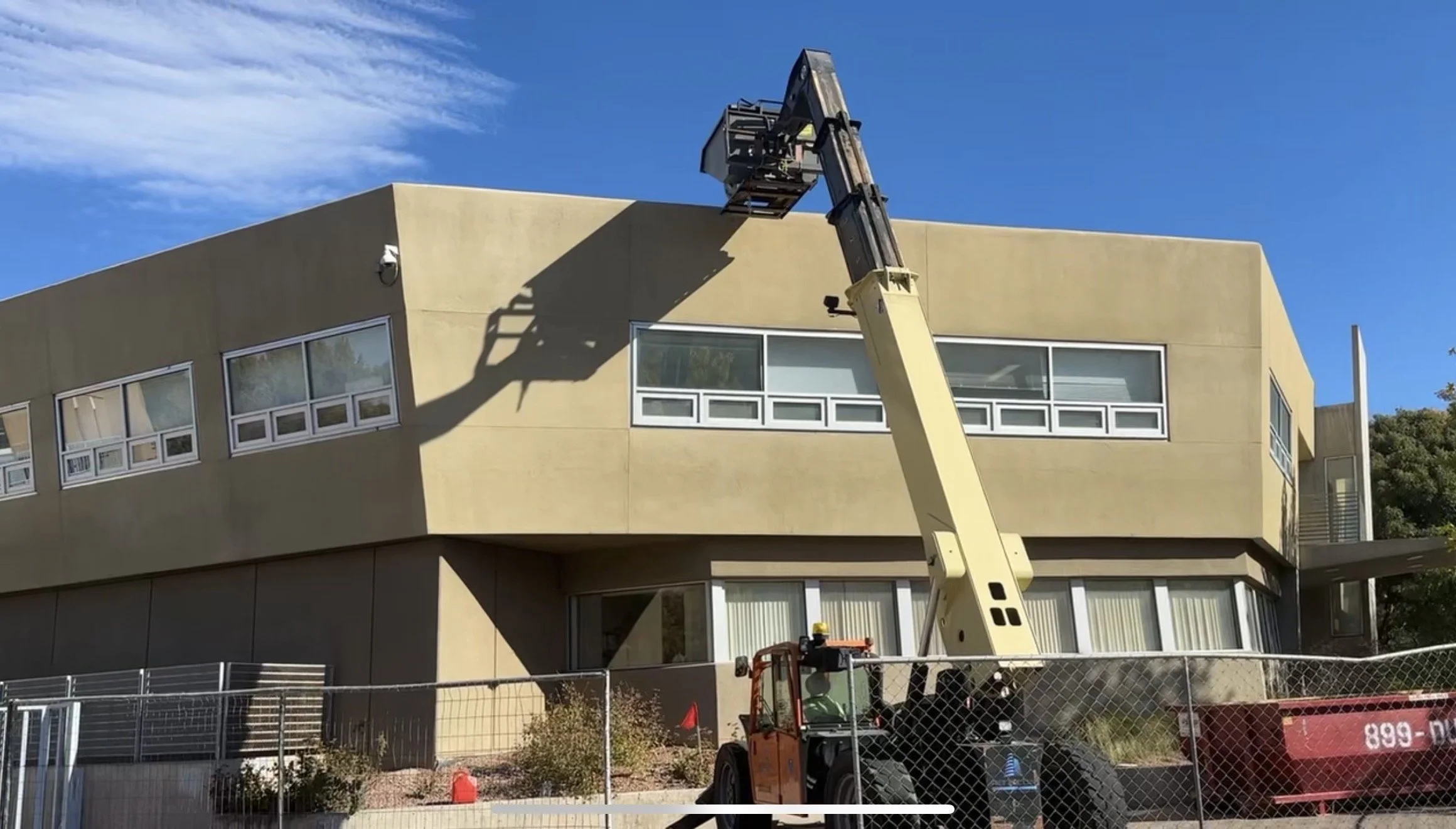 A construction or maintenance worker using an aerial lift to work on the exterior of a modern, multi-story building with large windows on a sunny day.