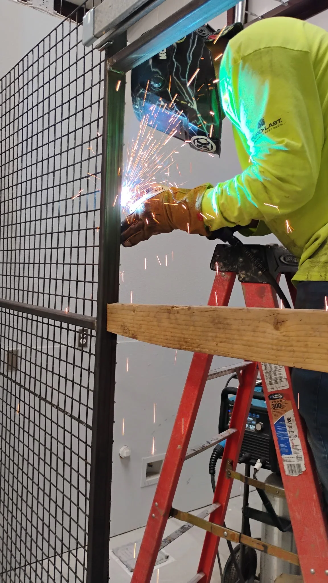 A person welding metal on a red ladder, wearing a yellow safety jacket and a welding helmet, sparks flying as they work on a metal frame.
