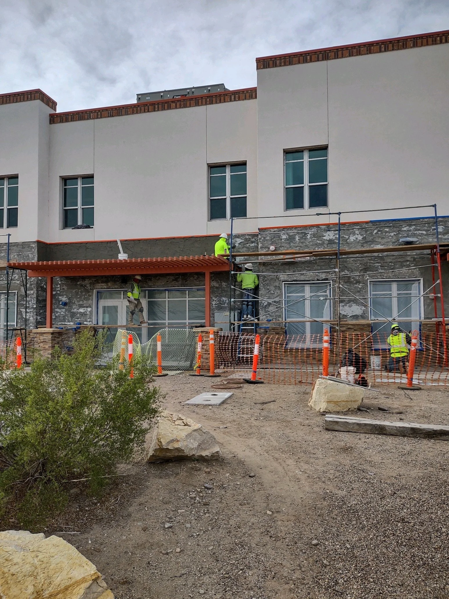 Construction workers wearing high-visibility jackets and helmets working on the exterior of a building, with scaffolding, orange safety fencing, and construction materials in the foreground.