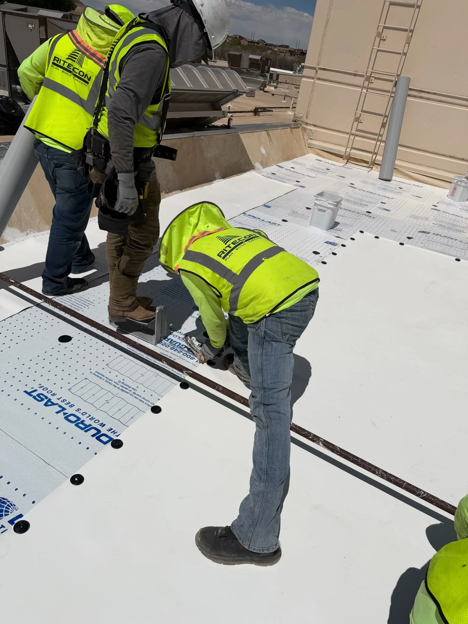 Workers on a rooftop installing large white panels, wearing neon yellow safety vests with company logo, tools around them, and blue sky in the background.