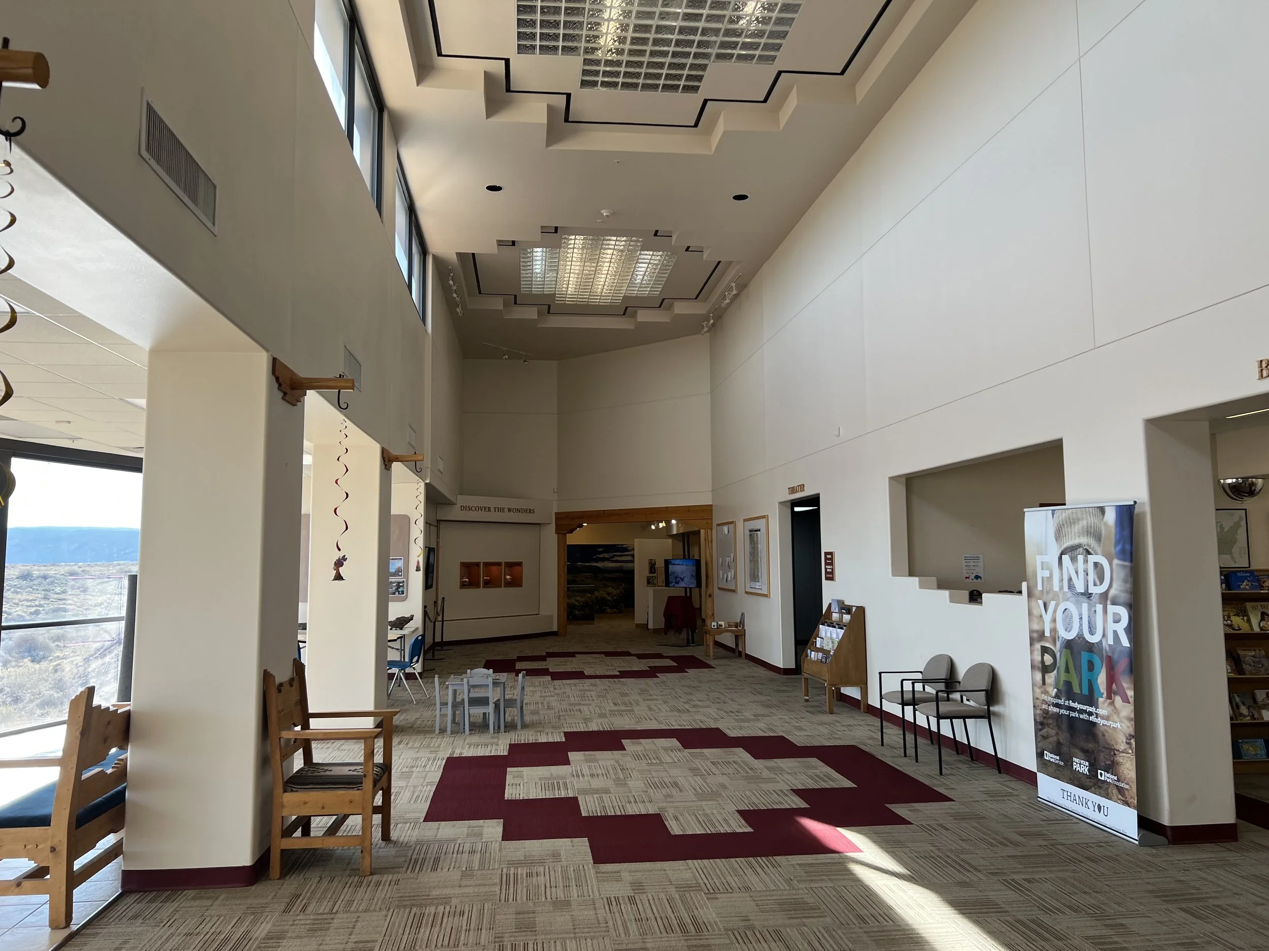 Interior of a spacious building with high ceilings, large windows, wooden chairs, and red and beige carpet flooring.