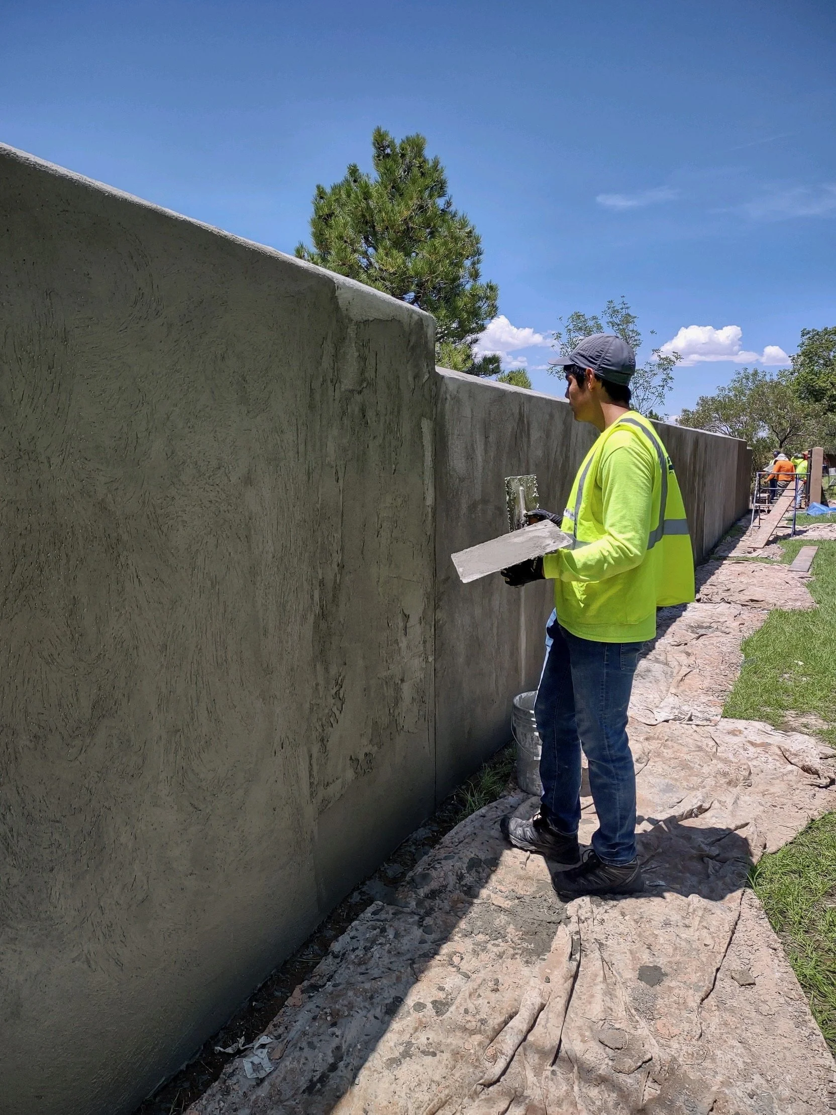 A construction worker in a bright yellow safety shirt and gray baseball cap stands beside a large, freshly poured concrete wall, with other workers visible in the background, under a clear blue sky with green trees surrounding the site.