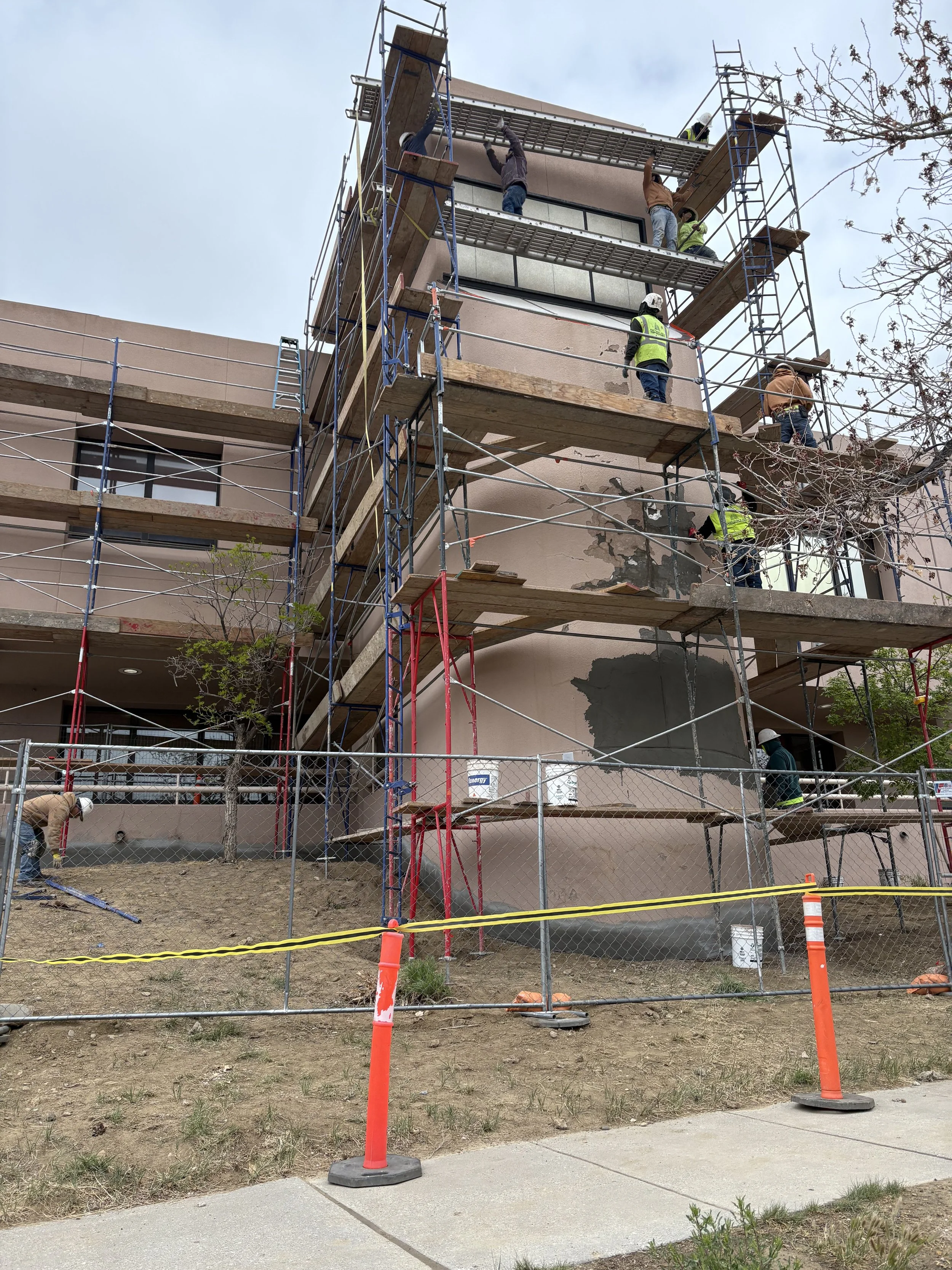 Multiple construction workers wearing safety helmets and vests working on scaffolding outside a building. The building’s exterior wall is being repaired with patching material. Safety cones and yellow caution tape cordon off the area.