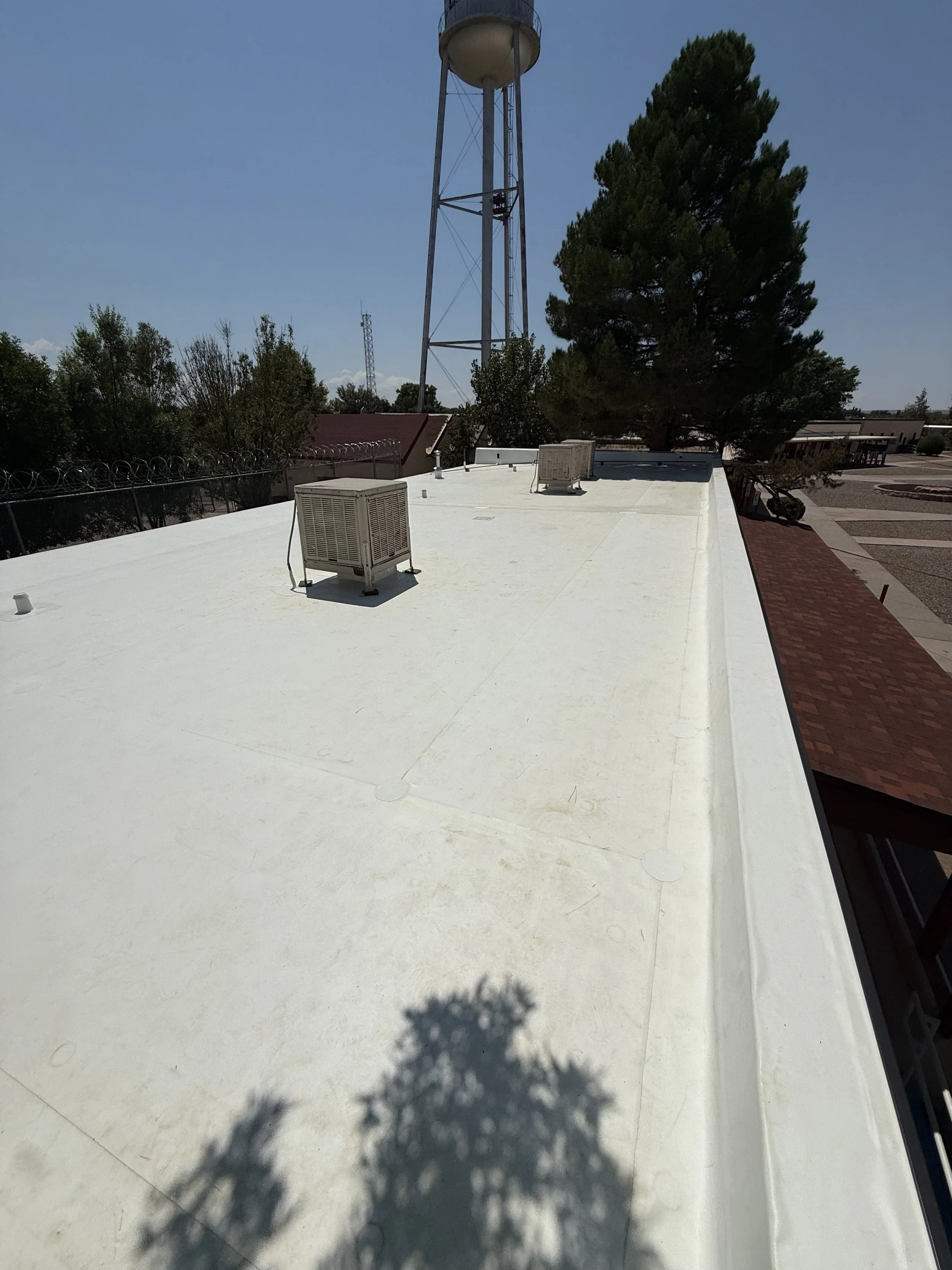 A flat rooftop with two air conditioning units, a water tower, several trees, and a building with a red roof under a clear blue sky.