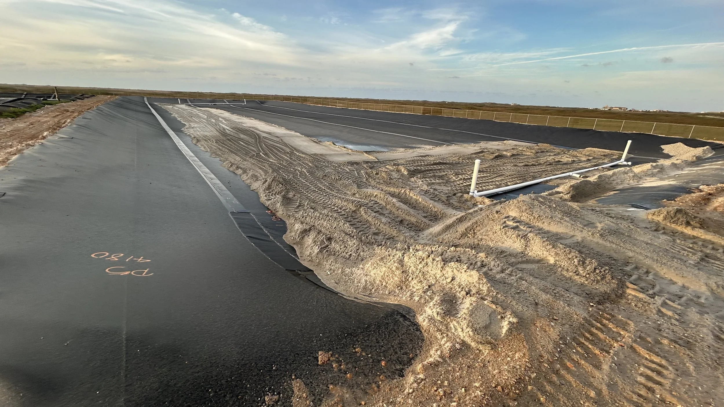 Construction site with partially paved road, disturbed dirt and sand, and exposed pipework, under a cloudy sky in a rural area.