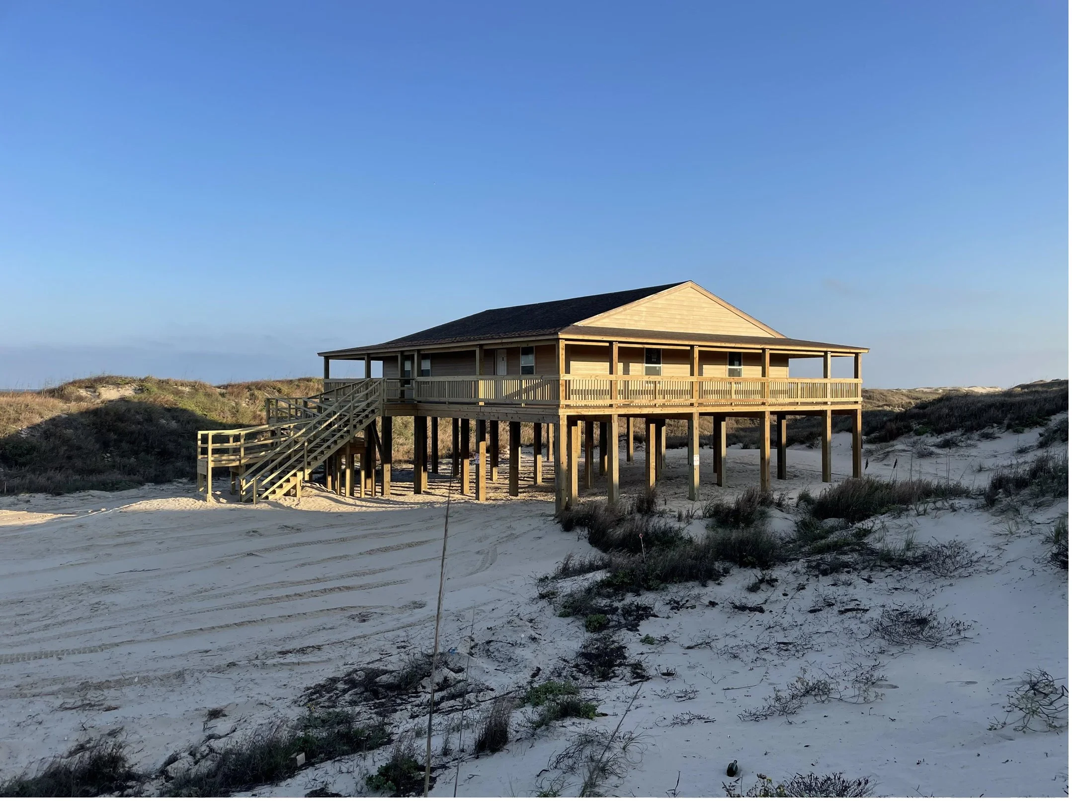 A wooden house on stilts with stairs leading up to a porch, set on a sandy area with sparse vegetation, under a clear blue sky.