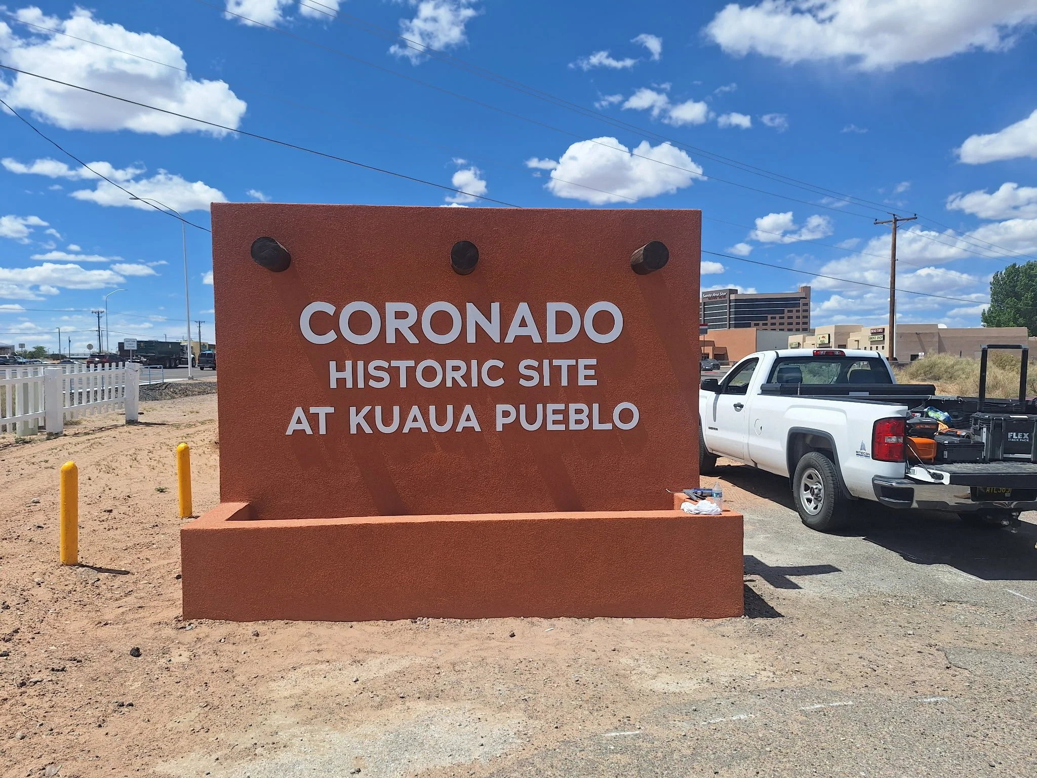 A red-orange sign reads 'Coronado Historic Site at Kuaua Pueblo,' with the sky and clouds in the background. There are cars, utility poles, and buildings surrounding the area.