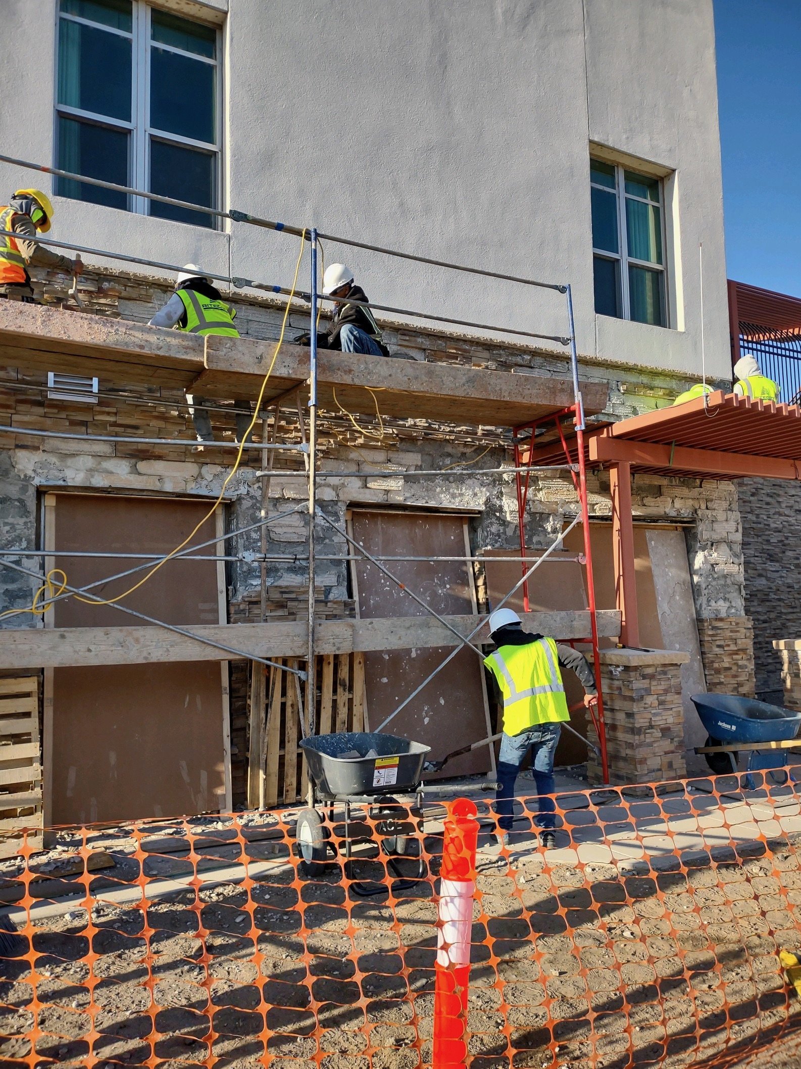 Construction workers on a building site, working on the exterior wall of a building, surrounded by orange safety fence and scaffolding, with windows on the building.