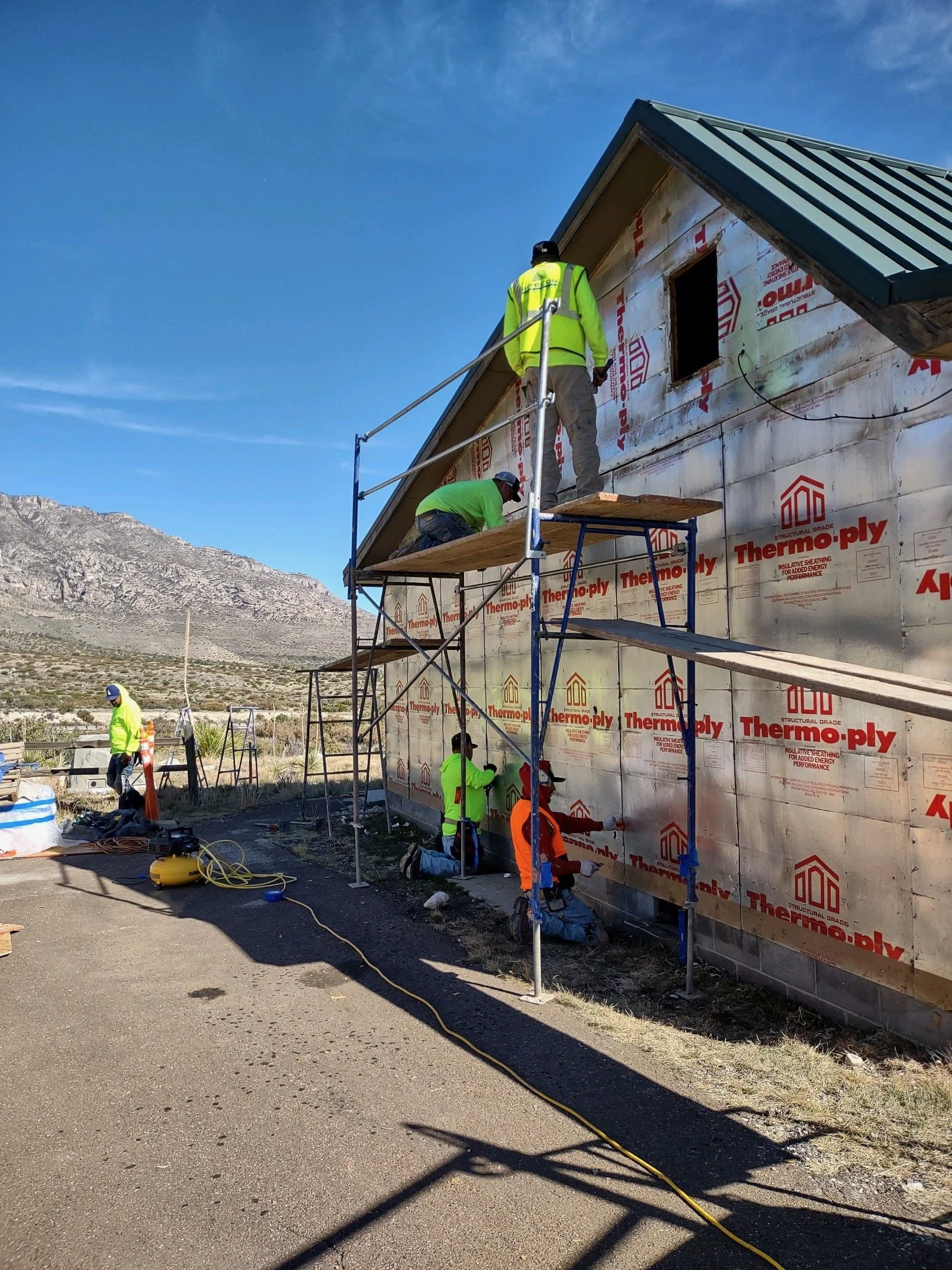 Construction workers building a house, with scaffolding and insulation on the exterior wall, in a desert landscape with mountains in the background.