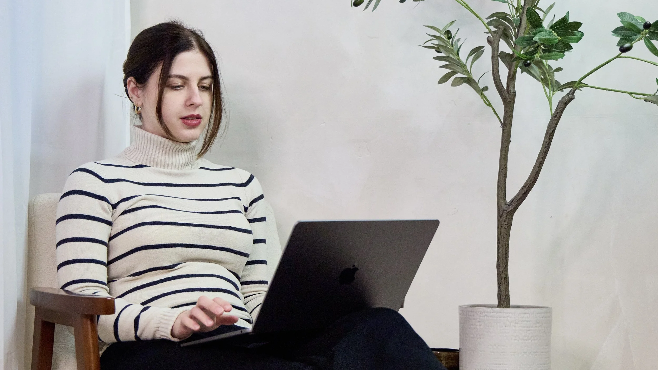 A woman with dark hair wearing a cream and navy striped turtleneck sweater sitting on a chair, using a silver laptop, with a tall potted plant beside her against a plain light-colored wall.