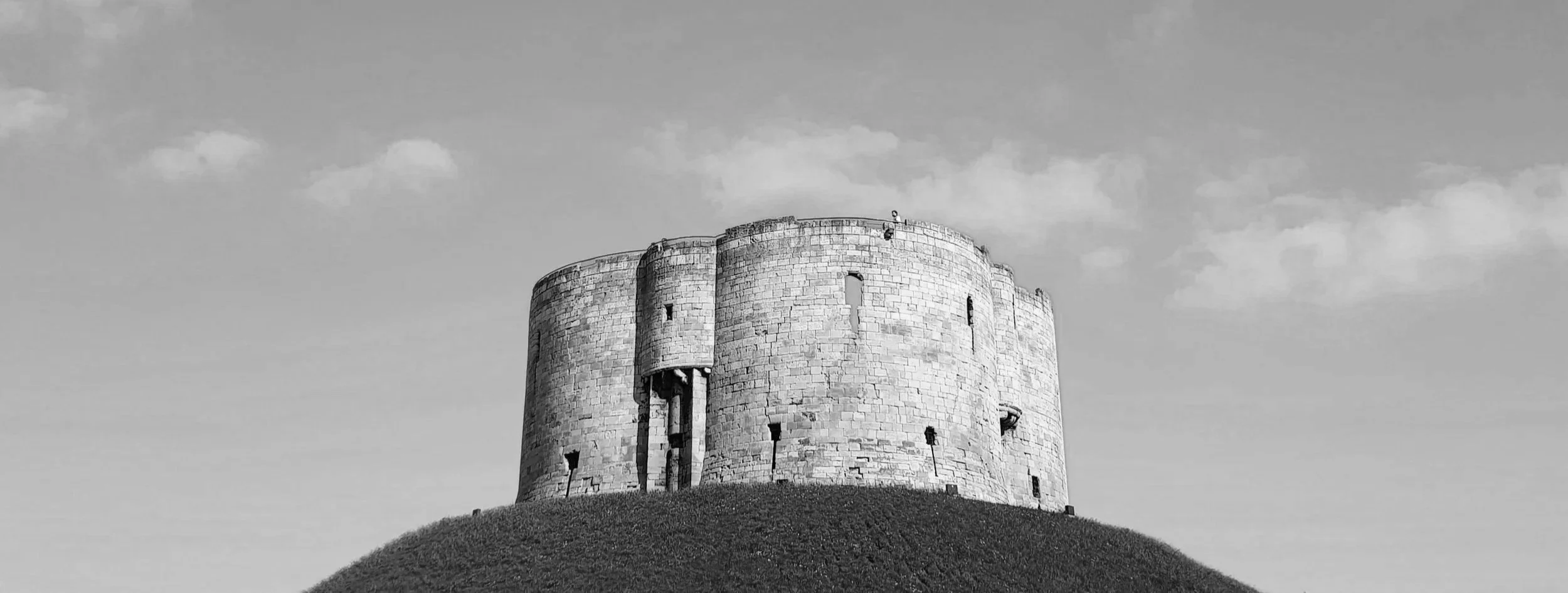 A black and white photo of an old castle or fort on a grassy hilltop, with a partly cloudy sky in the background.