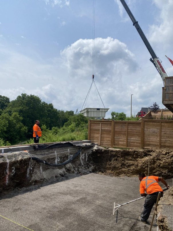 Construction workers in orange safety shirts leveling a gravel area at a construction site with a crane lifting a large container in the background.