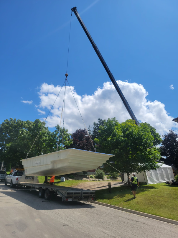 A crane lifting a large white boat off a trailer on the street, with workers in safety vests nearby and trees in the background under a partly cloudy sky.