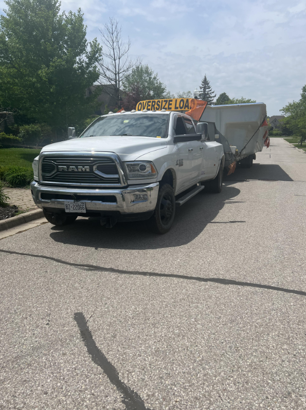 A white RAM pickup truck with an orange 'Oversize Load' sign on top, attached to a large gray trailer with a rusty brown cover, parked on a suburban street under a partly cloudy sky.