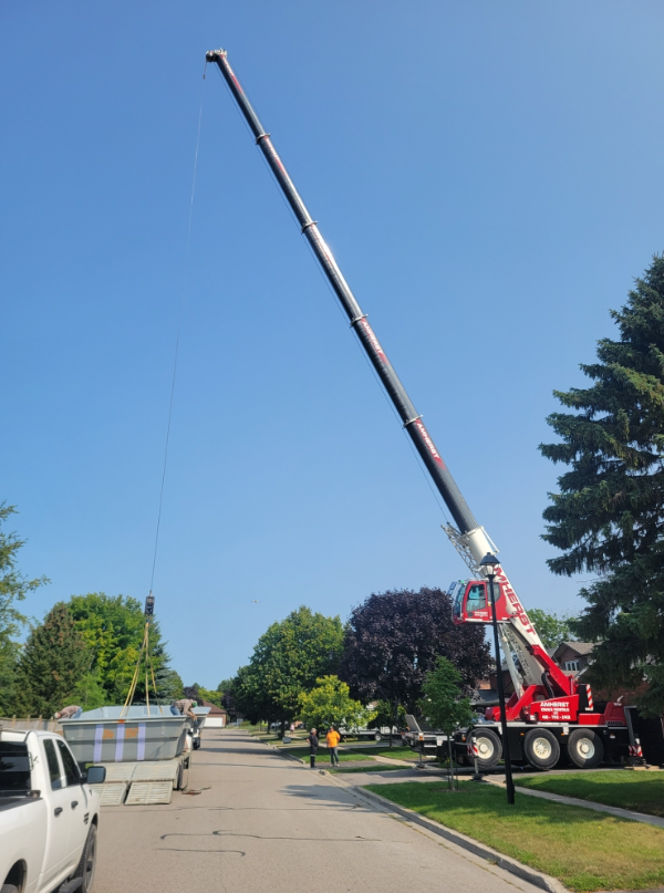 A large crane lifting a hot tub over a residential street with trees and parked cars.