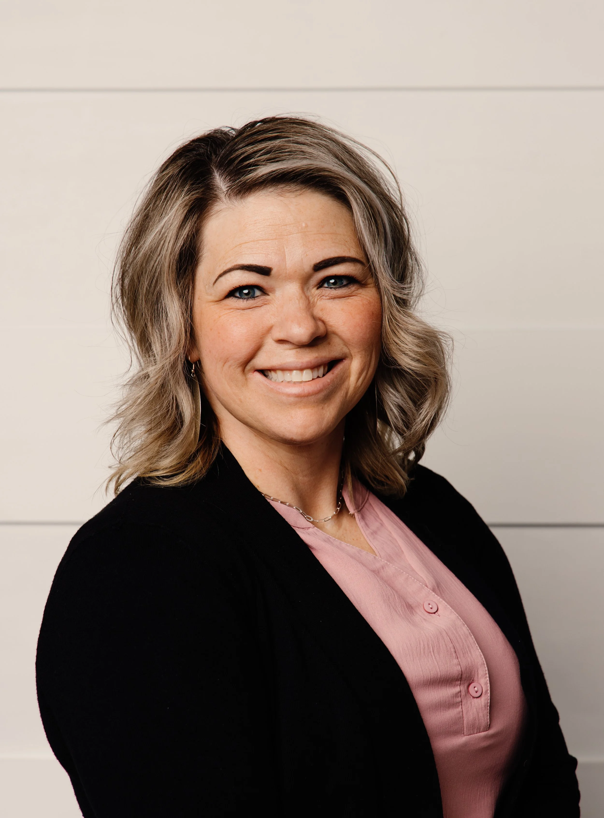 A woman with shoulder-length wavy gray hair and blue eyes smiling, wearing a black blazer over a pink button-up shirt, standing against a white wall.