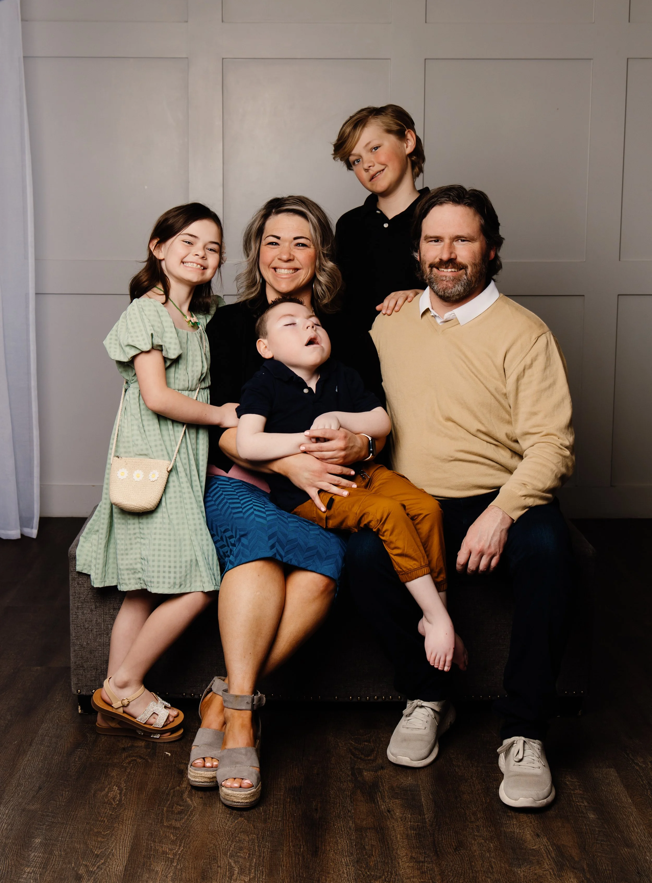 Family portrait with three children and two adults, all smiling, indoors against a wall with wooden flooring.