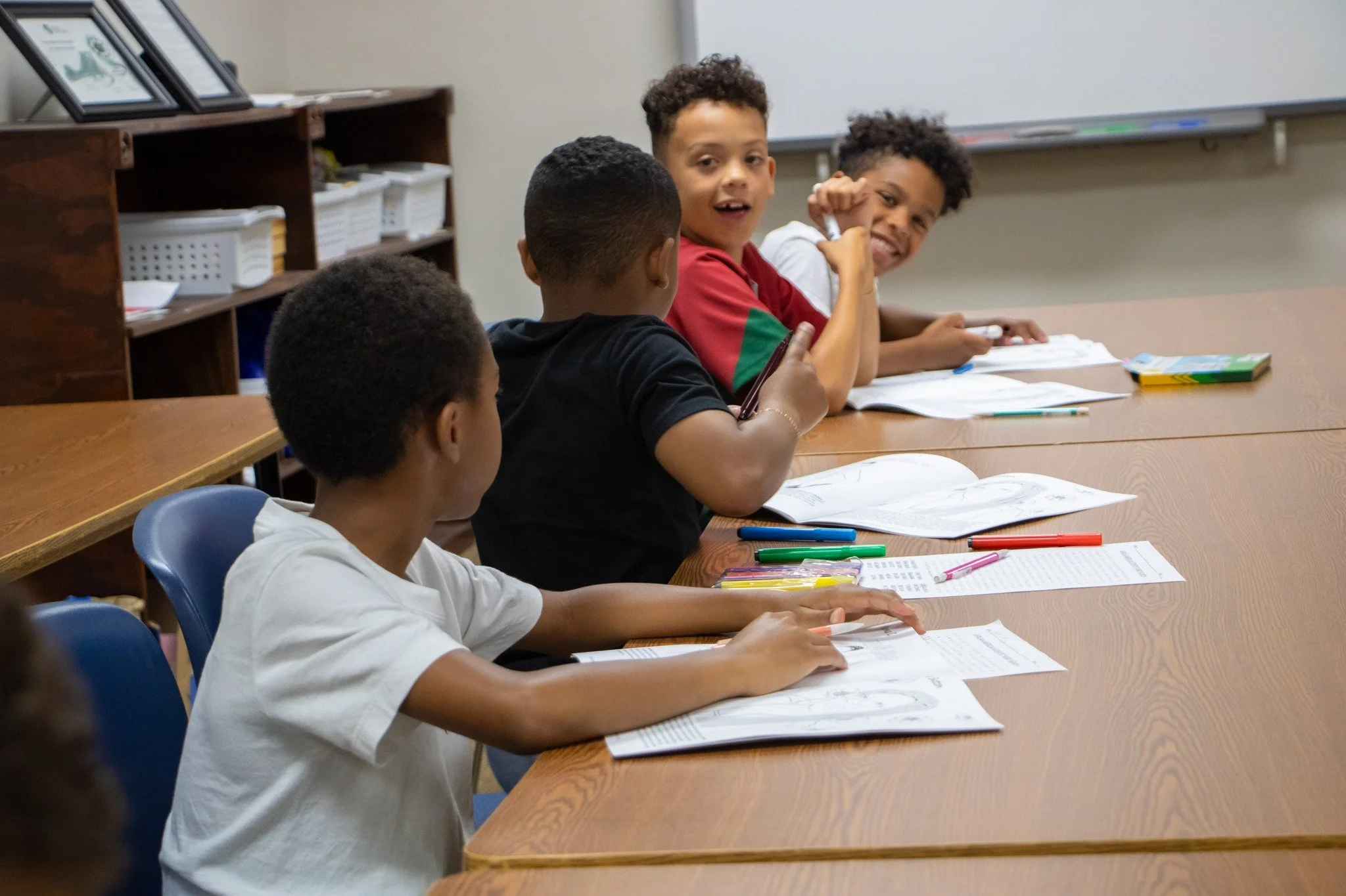 A classroom scene with four young boys sitting at a wooden table, engaged in learning activities with open textbooks, colored pens, and pencils, one boy smiling and looking at the camera.