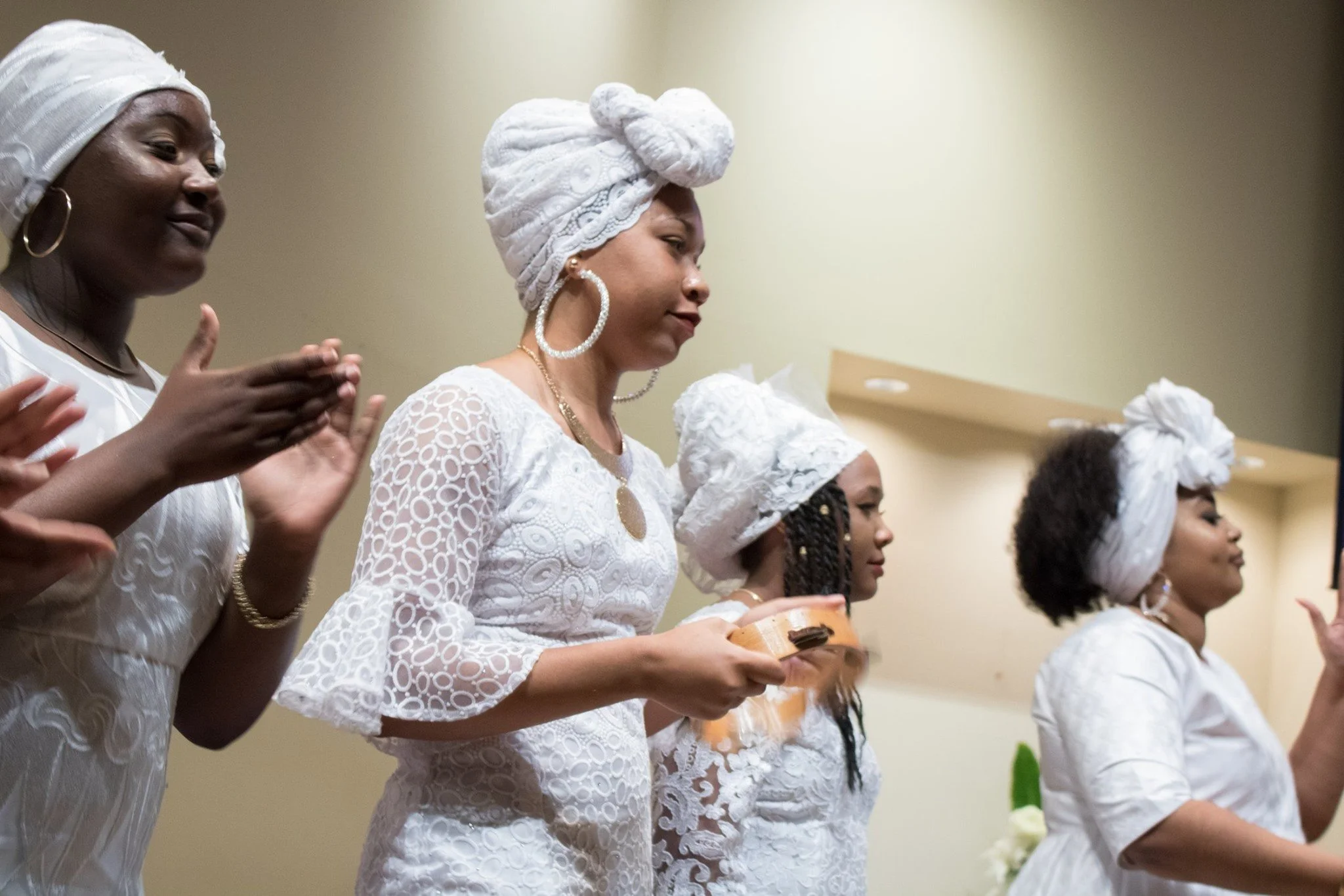 A group of women dressed in white, participating in a cultural or religious ceremony, with some wearing head wraps and large hoop earrings, and holding bowls or instruments.