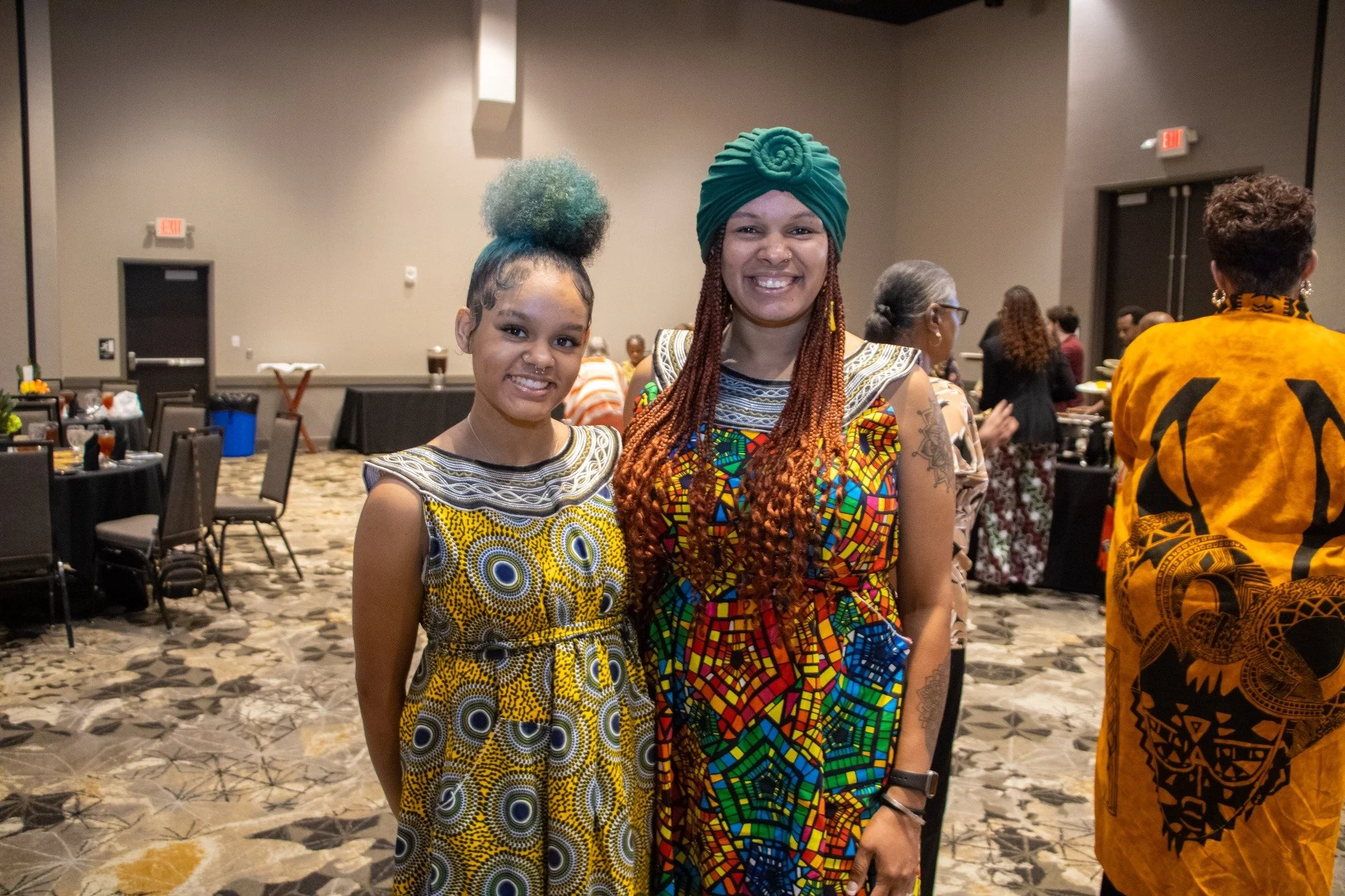 Two women wearing colorful African-inspired clothing smile at the camera, standing in a large room with multiple people and tables in the background.