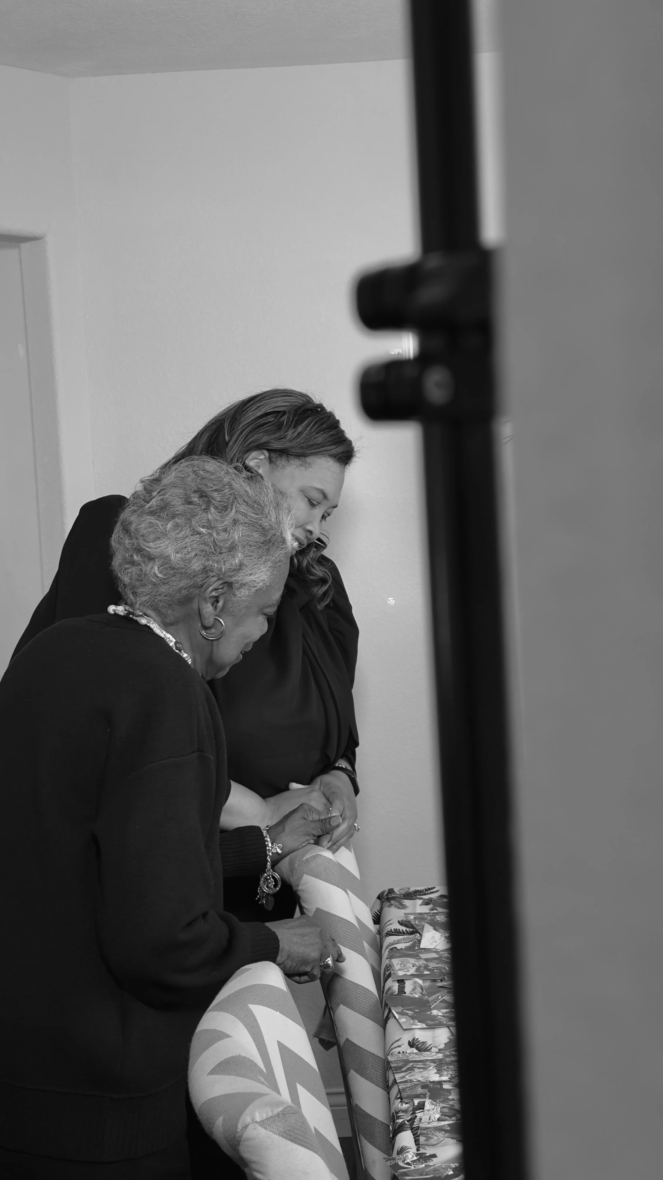 Black and white photo of two women, one older and one younger, standing close together and looking down at a table with gift-wrapped presents, partially obscured by a vertical object in the foreground.