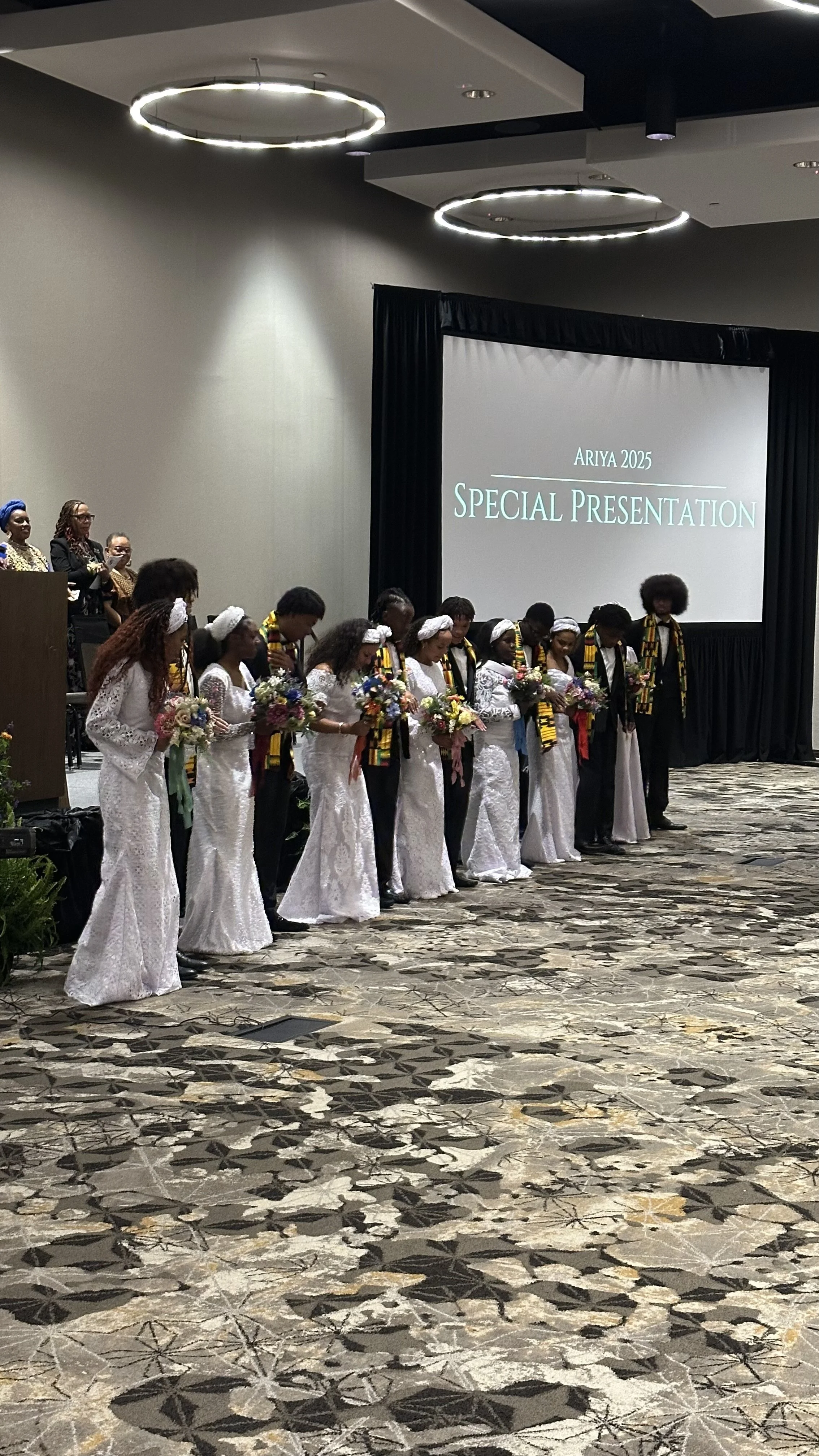 People dressed in white and black standing in a line holding flowers during a special presentation at an event with a large screen in the background displaying the text 'Ariya 2025 Special Presentation'.